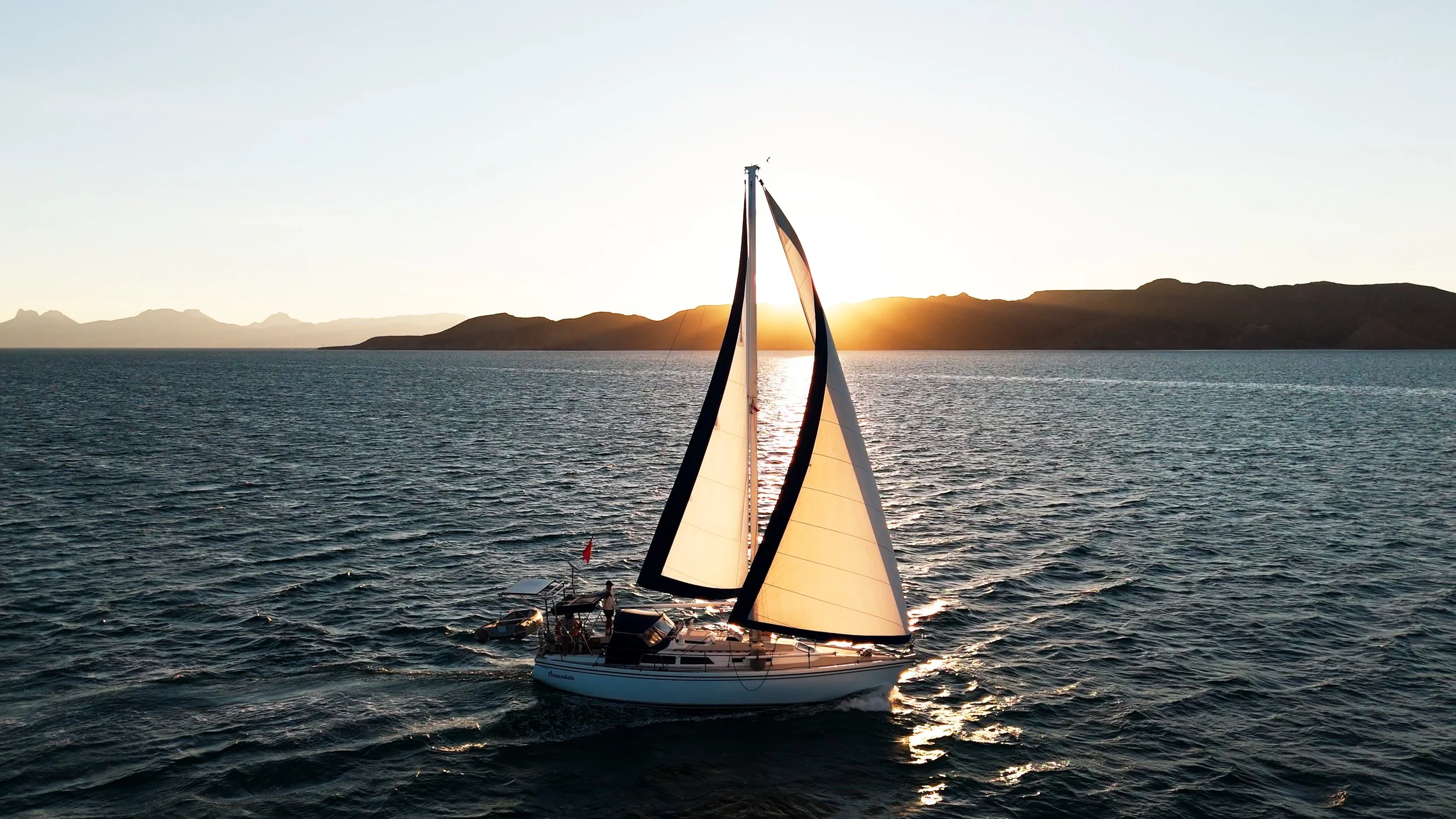 A sailboat with its sails up on the water during sunset, with mountains in the background.