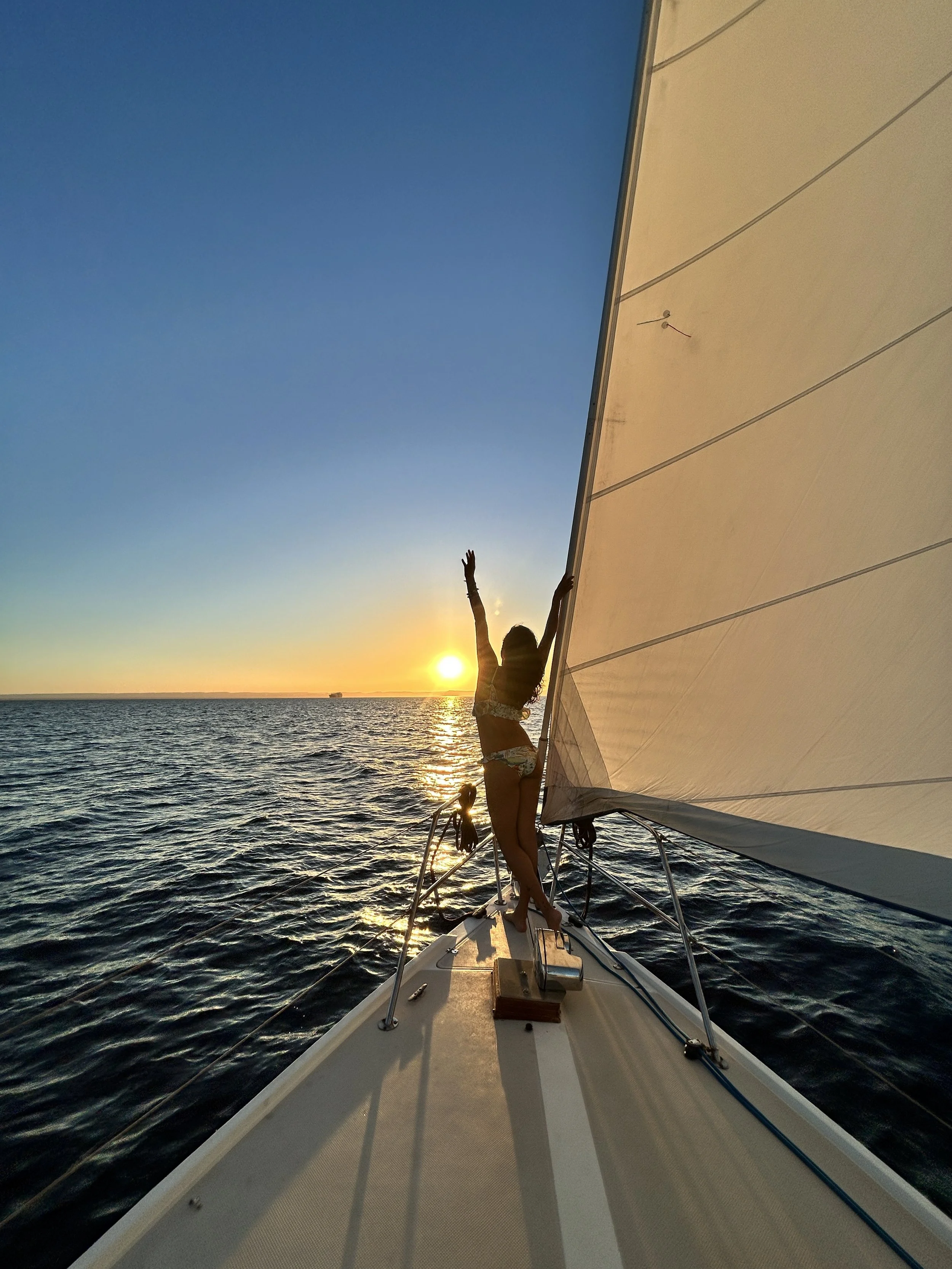 A woman standing on a sailboat at sunset, holding the sail with one hand and raising her other arm, with the ocean and a distant ship in the background.