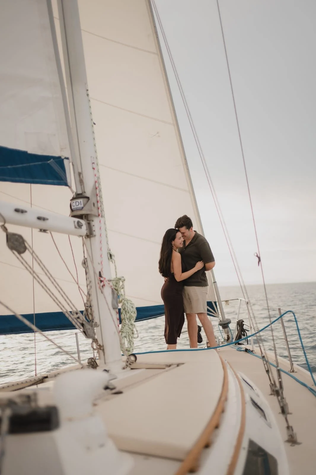 A couple embracing on a sailboat at sea, with the large sail and rigging visible in the foreground.