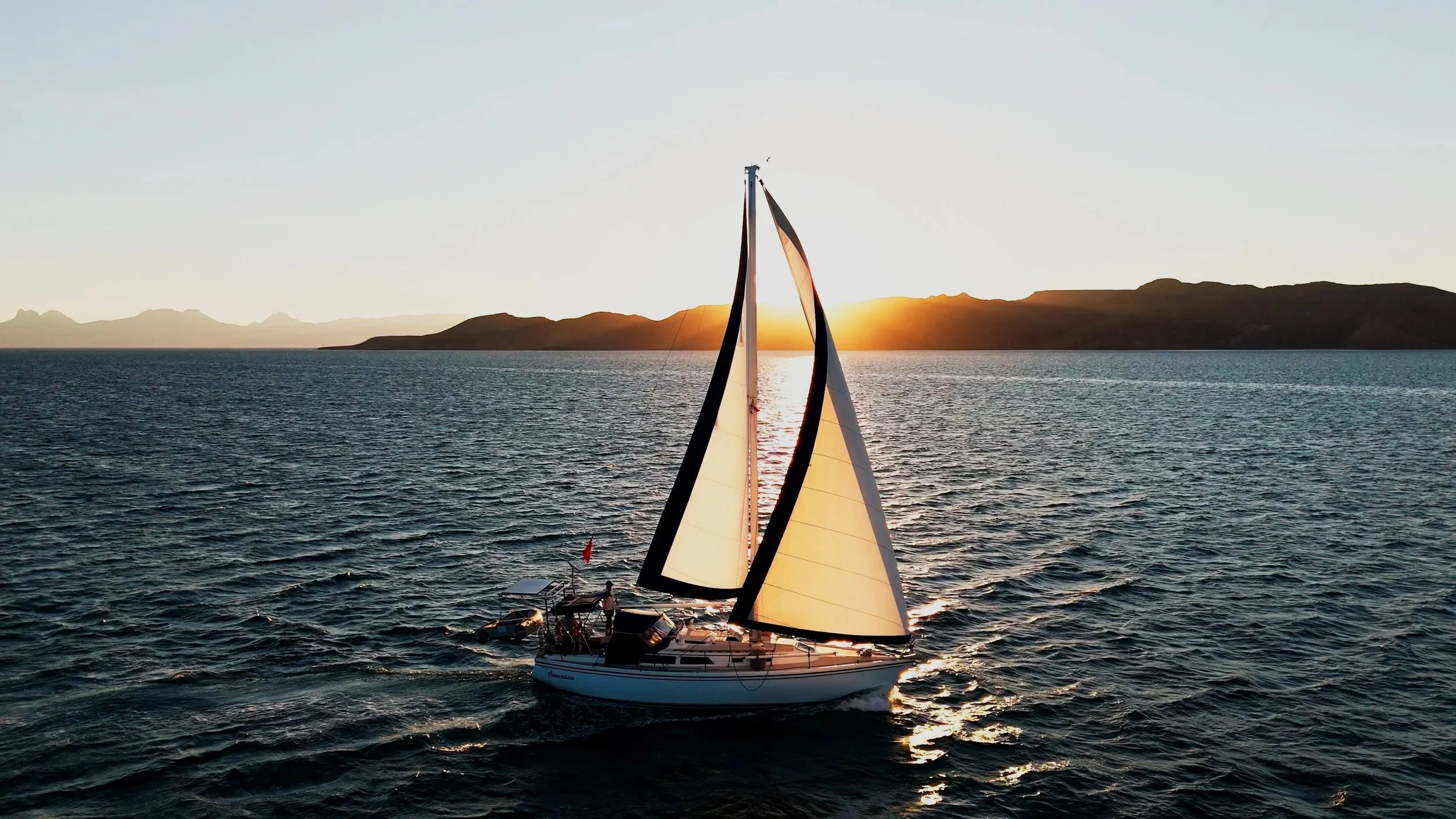 A sailboat on the ocean at sunset with a mountainous shoreline in the background.