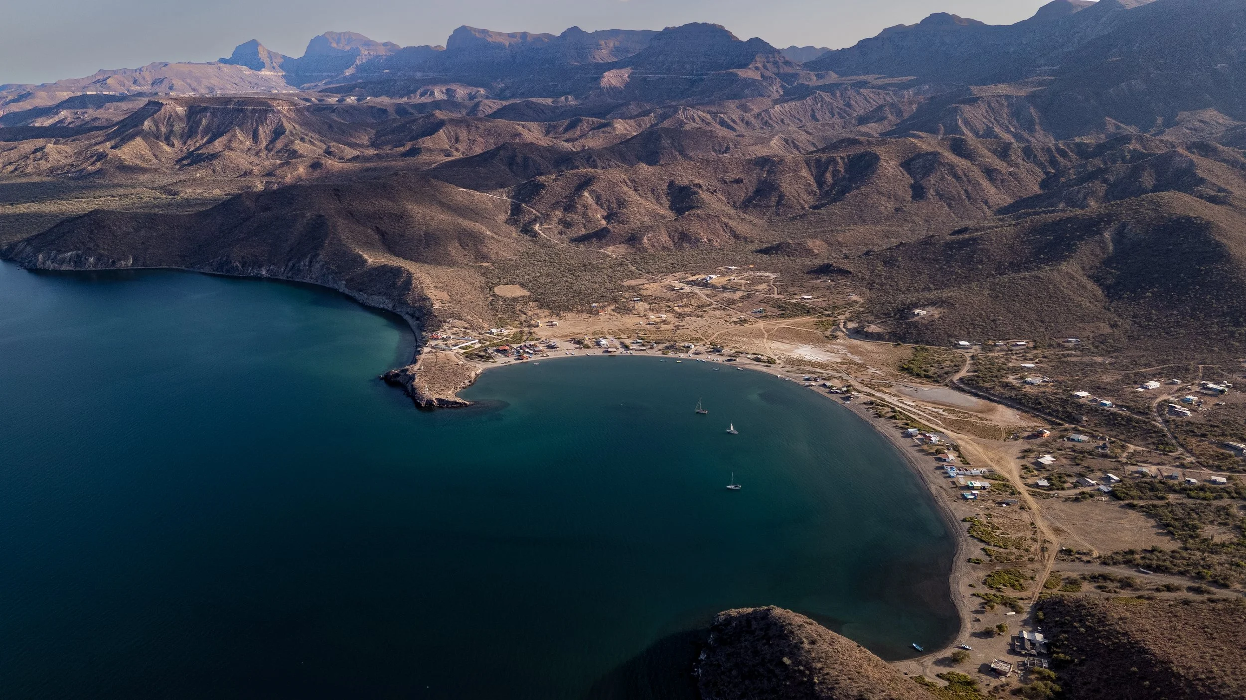 Aerial view of a desert landscape with a small bay, boats, and a shoreline with houses surrounded by hills and mountains.