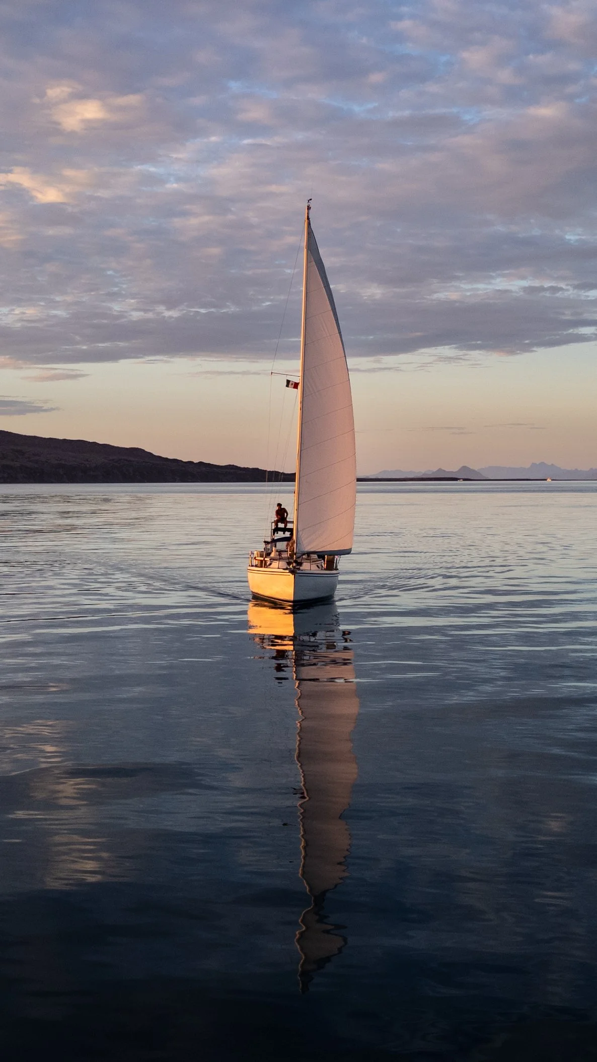 A sailboat with a single large white sail on calm water during sunset or sunrise. The sky has scattered clouds, and there is a person sitting on the boat. Distant land and mountains are visible in the background.