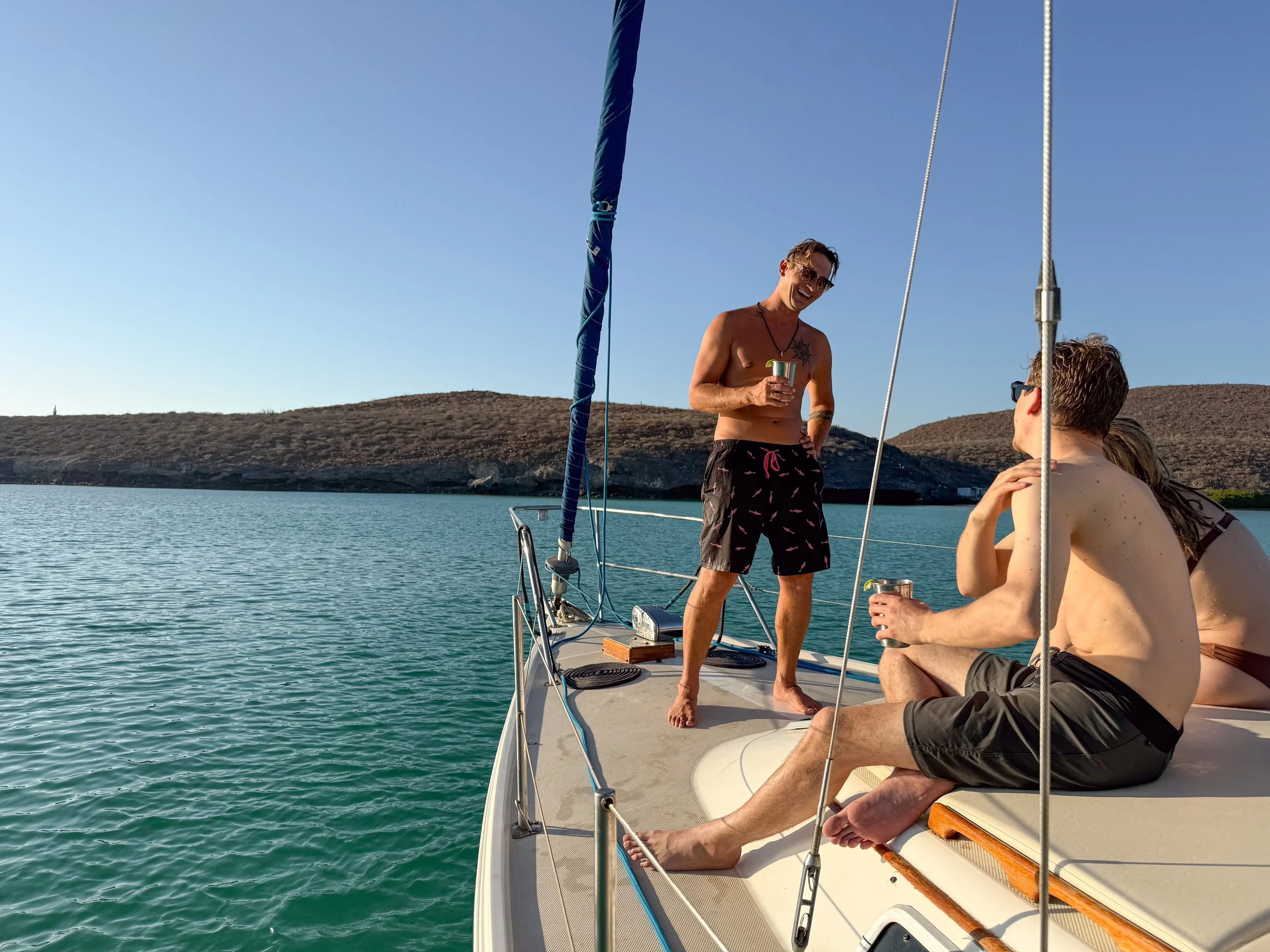 Three friends relaxing on a sailboat on a calm body of water, with dry hills in the background. One man is standing and talking, holding a drink, while the other two are sitting and listening, also holding drinks. The man standing is shirtless and wearing sunglasses and swim shorts.