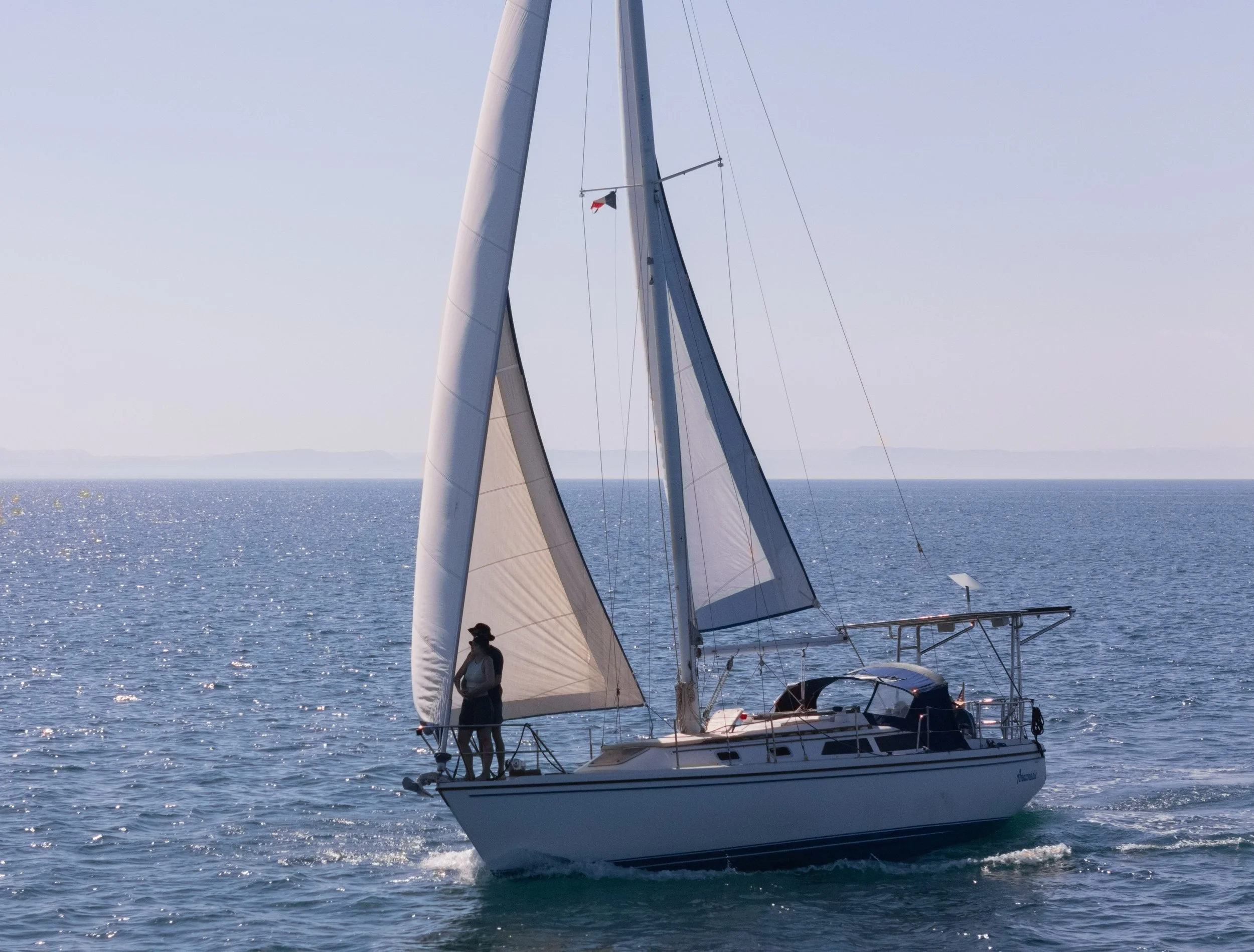 A sailboat with large white sails on the ocean with two people on board, under a clear sky with faint distant land in the background.