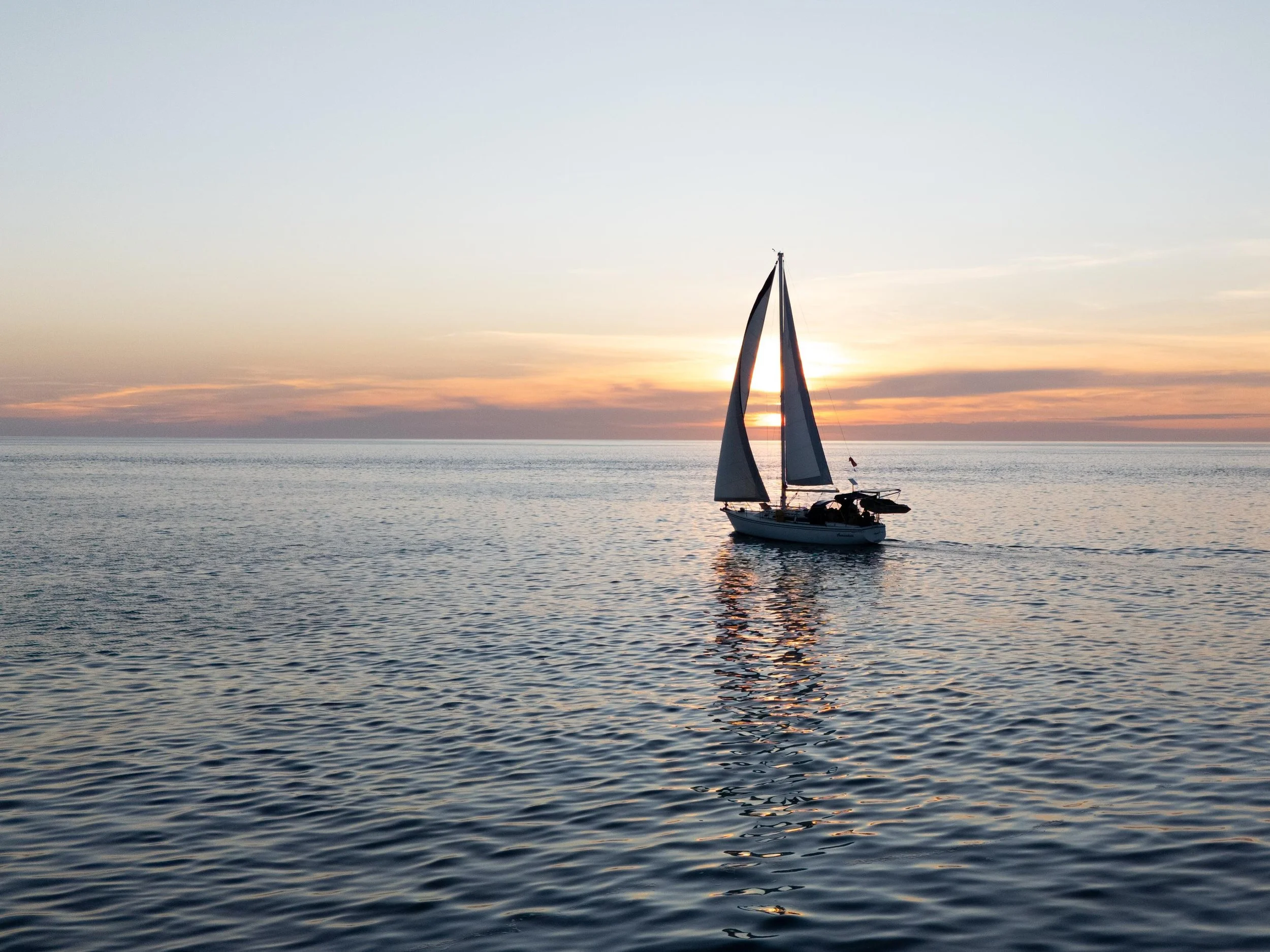 A sailboat with dark sails on calm water during sunset, with the sky and horizon visible in the background.