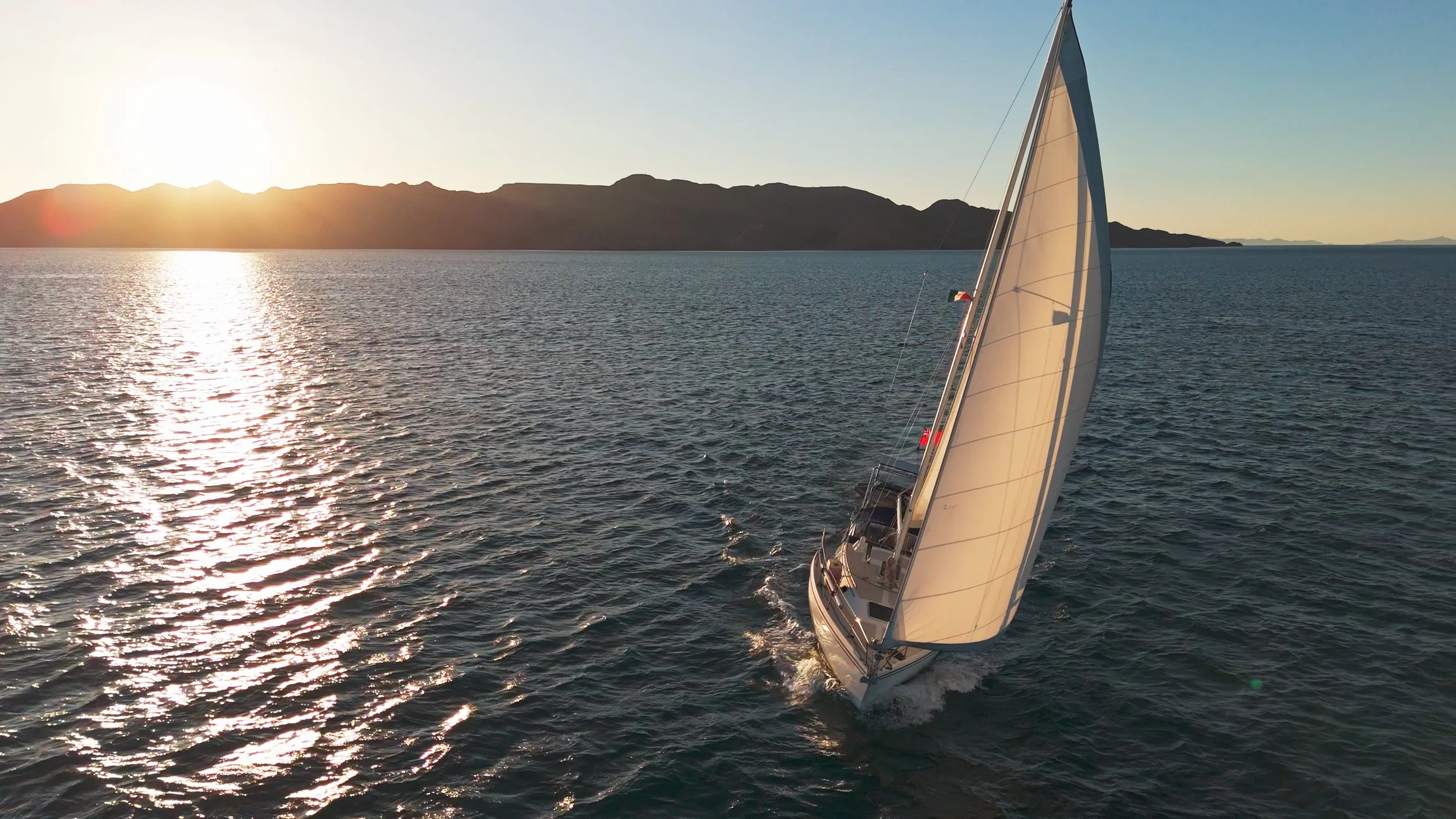 A sailboat on the ocean during sunset, with a mountain range in the background and the sun low on the horizon.