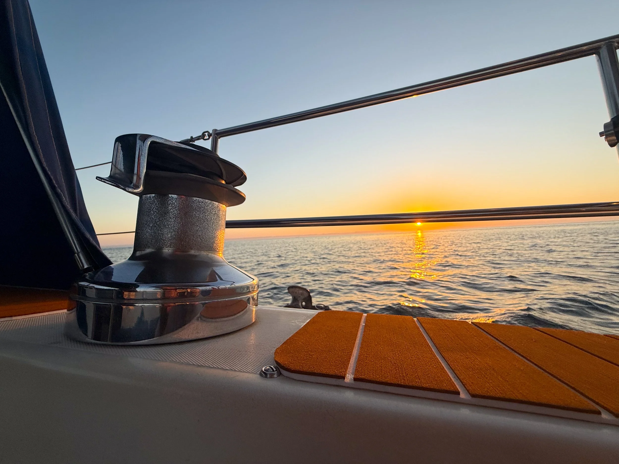 A yacht's deck at sunset with a metal winch, wooden planks, and a view of the calm sea and sunset in the distance.