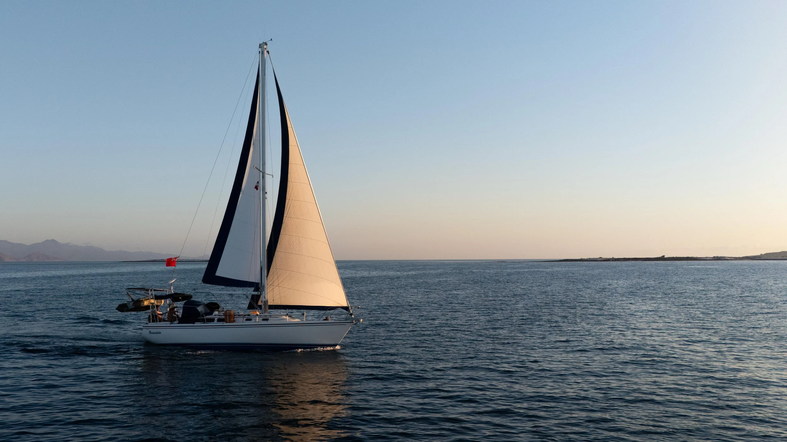 A sailboat with white sails and a tan sail on calm blue water during sunset, with land and mountains in the distance.