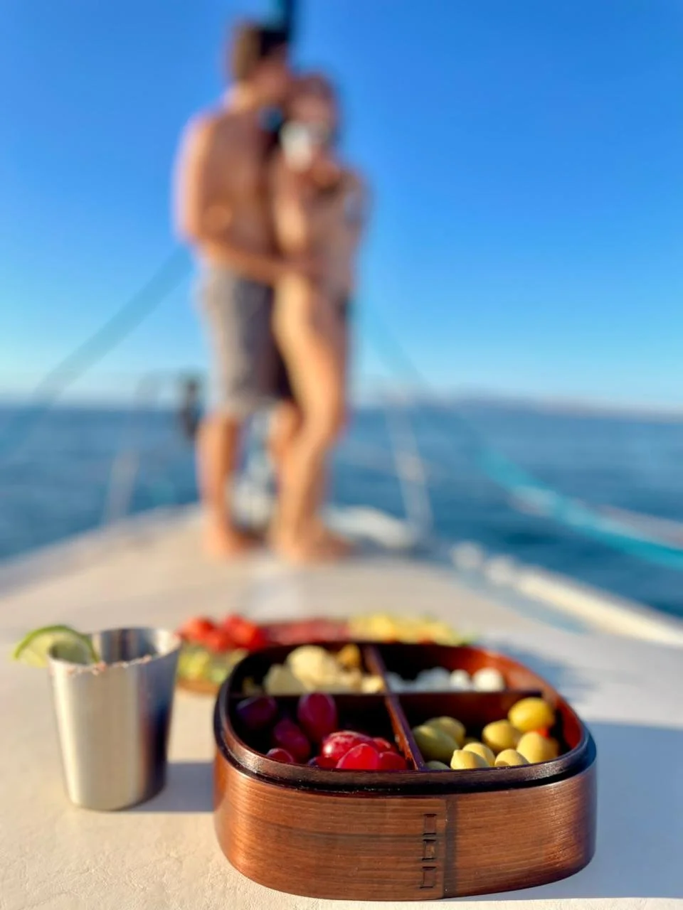 Close-up of a wooden divided snack box filled with grapes and other snacks on a boat, with a blurred couple taking a selfie in the background.