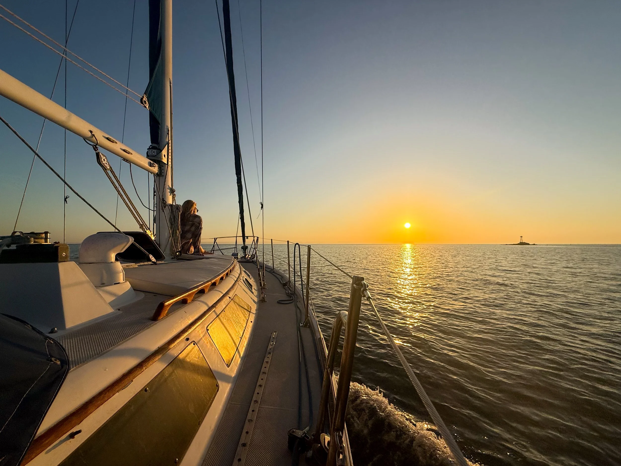 A person sitting on the deck of a sailboat during sunset, with the ocean and a lighthouse in the distance.