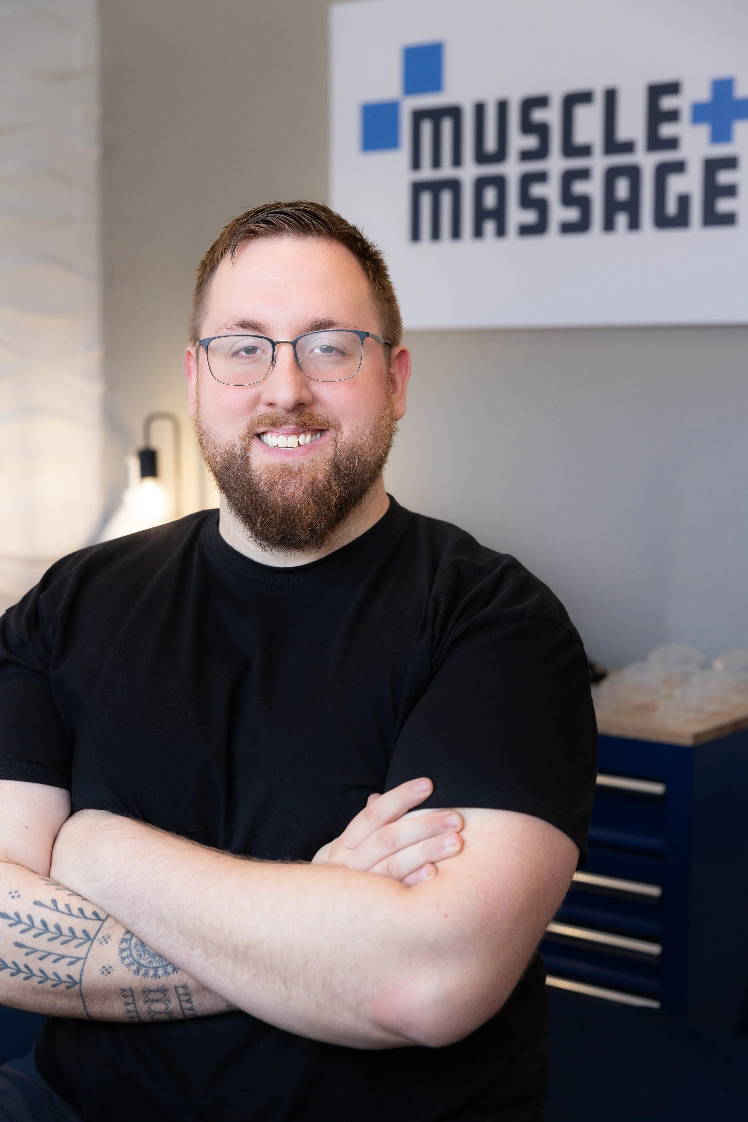 A smiling man with glasses and a beard, wearing a black t-shirt, standing with crossed arms in front of a sign that reads 'Muscle + Massage' on a white wall.