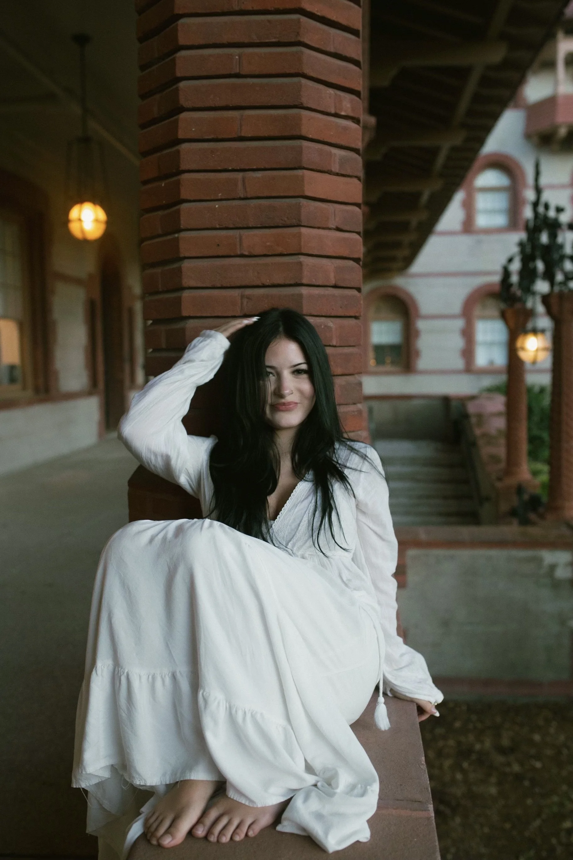 A woman with black hair and light makeup sitting on a bench outside at dusk, wearing a loose white dress with long sleeves and tassel details, leaning her head on a brick pillar with a slight smile. In the background, there are buildings with arched windows and decorative trim, and warm glowing lights hanging from the building.