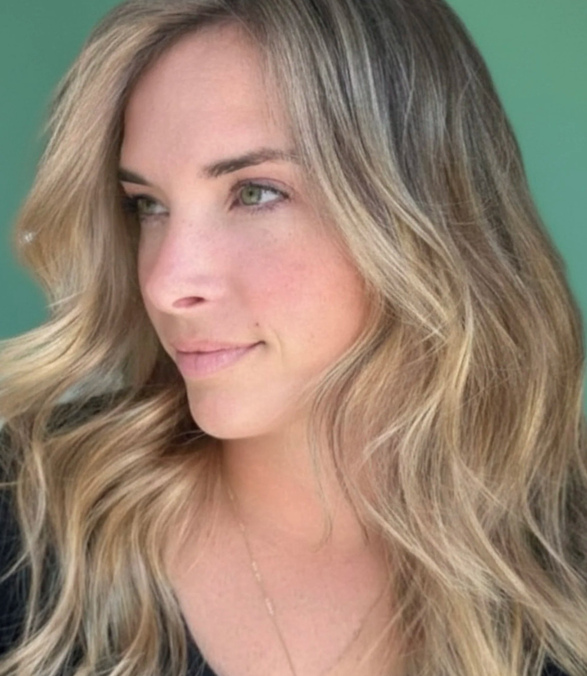 Close-up of a woman with blonde, wavy hair and light skin, smiling softly, with a portion of her eye and forehead visible against a green background.