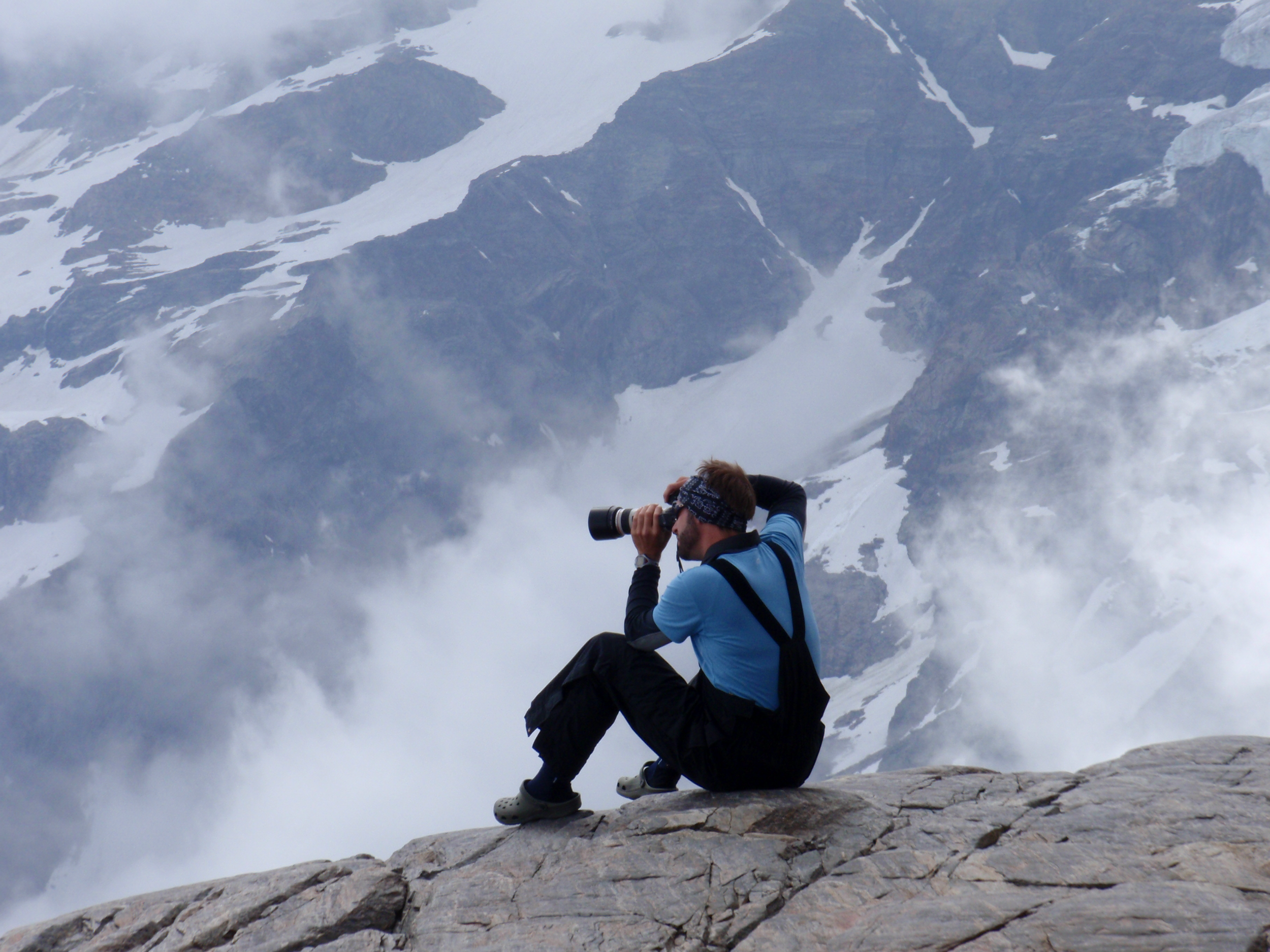 Person som sitter på en bergstopp med en kamera, omgiven av snötäckta berg och moln.