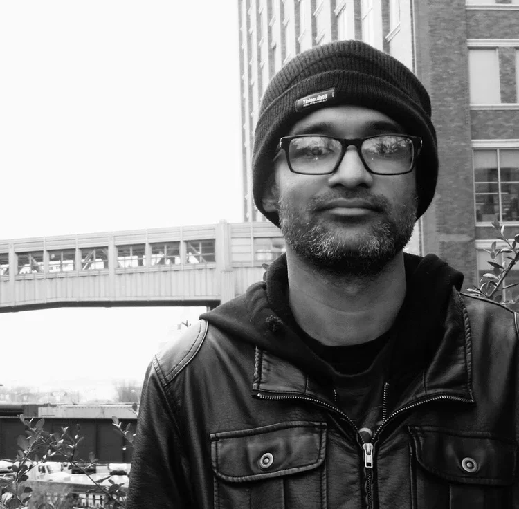 Man wearing glasses, beanie, and leather jacket standing outdoors in an urban setting.