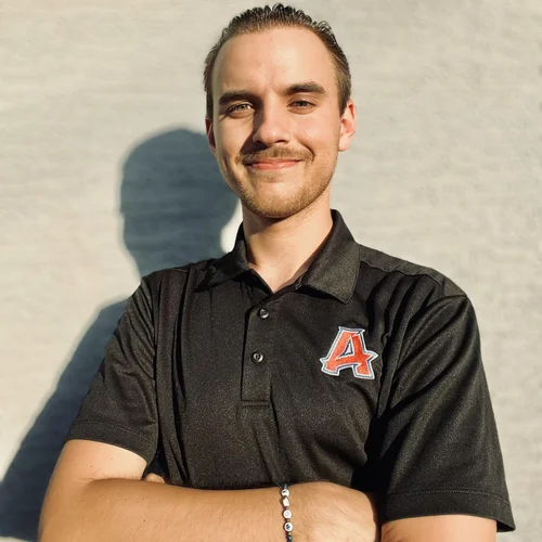 A young man with light skin, dark hair, and a beard, smiling with arms crossed, wearing a black polo shirt with an orange and white letter A on the chest, standing outdoors in front of a whitish wall with a shadow behind him.