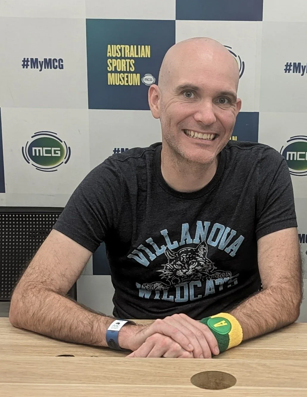 A smiling man with a shaved head sitting at a table in front of a backdrop with Australian Sports Museum and MCG logos. He wears a display wristband, a black T-shirt with a Villanova Wildcats logo, and a green and yellow wristband.