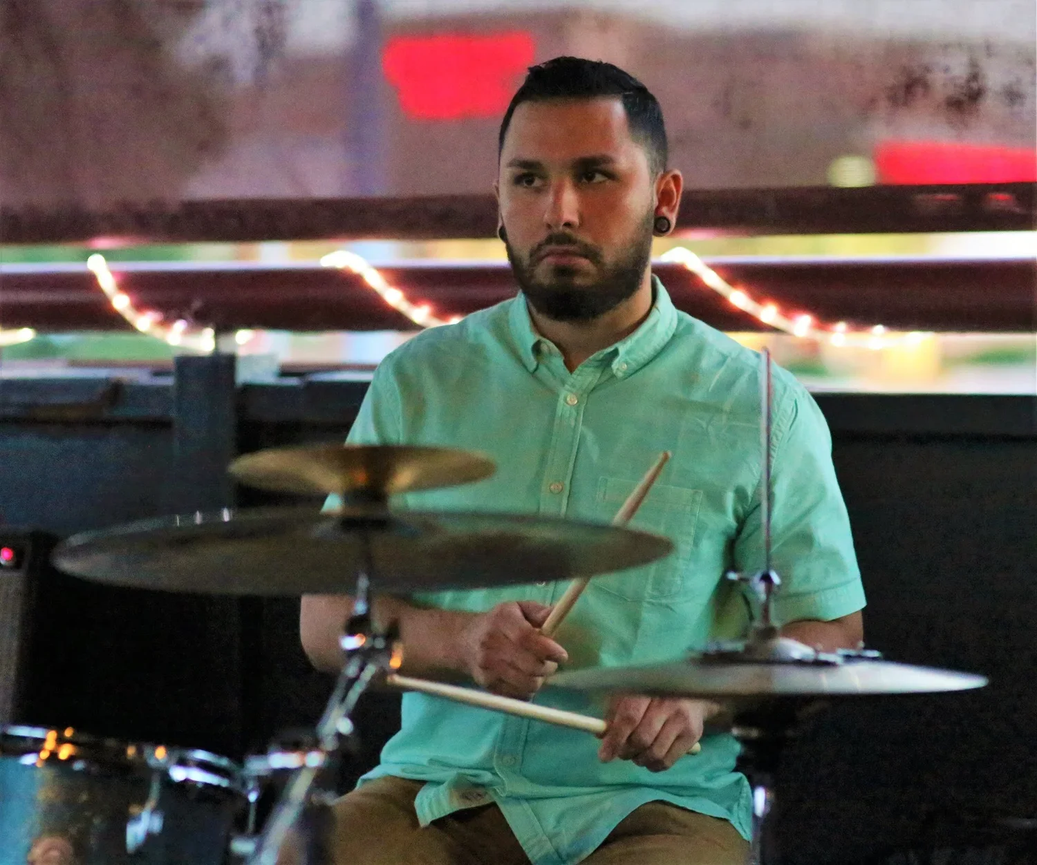 A man with a beard and earrings playing drums in a dimly lit setting with colorful fairy lights in the background.