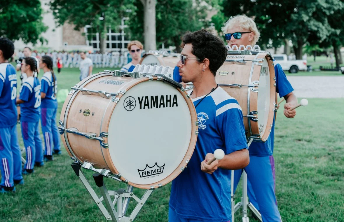 Young man and woman drummers in blue uniforms playing drums outdoors at a community event, with people standing in line in the background and trees nearby.