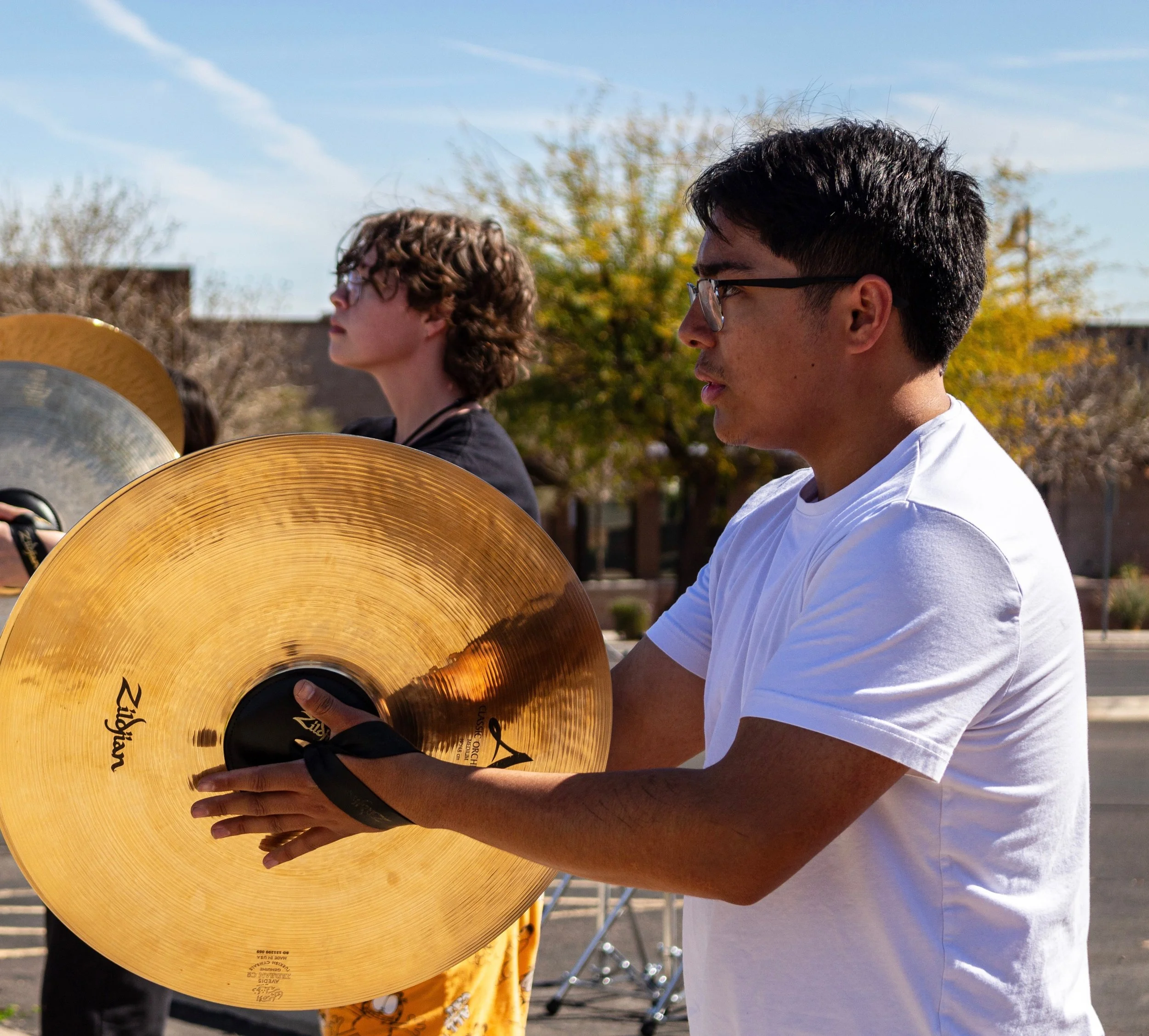 A young man in a white t-shirt and glasses playing a cymbal outdoors, with other musicians and trees in the background.