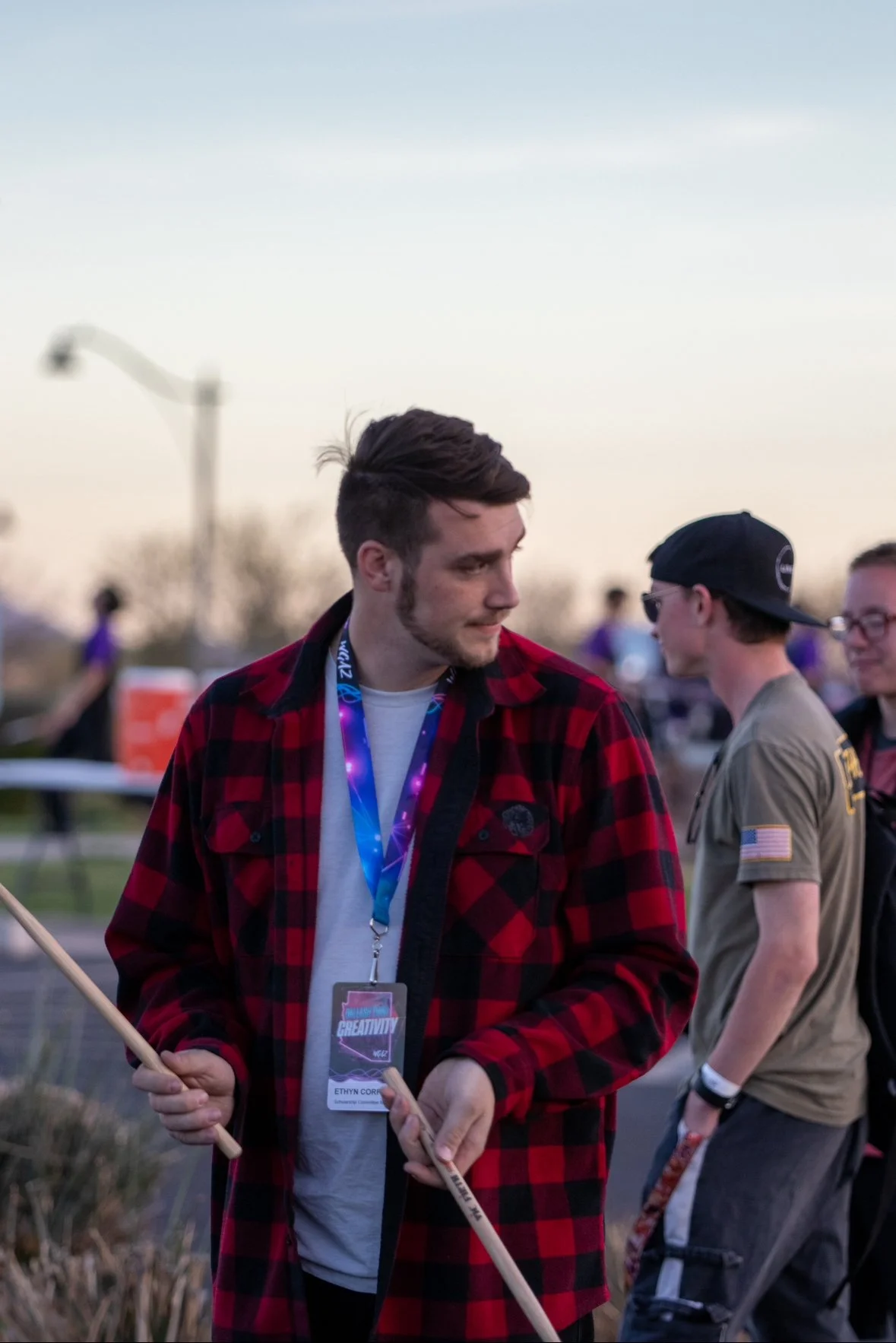 A young man with brown hair, a beard, and a red and black plaid shirt looking down while holding a wooden stick during outdoor event at dusk.