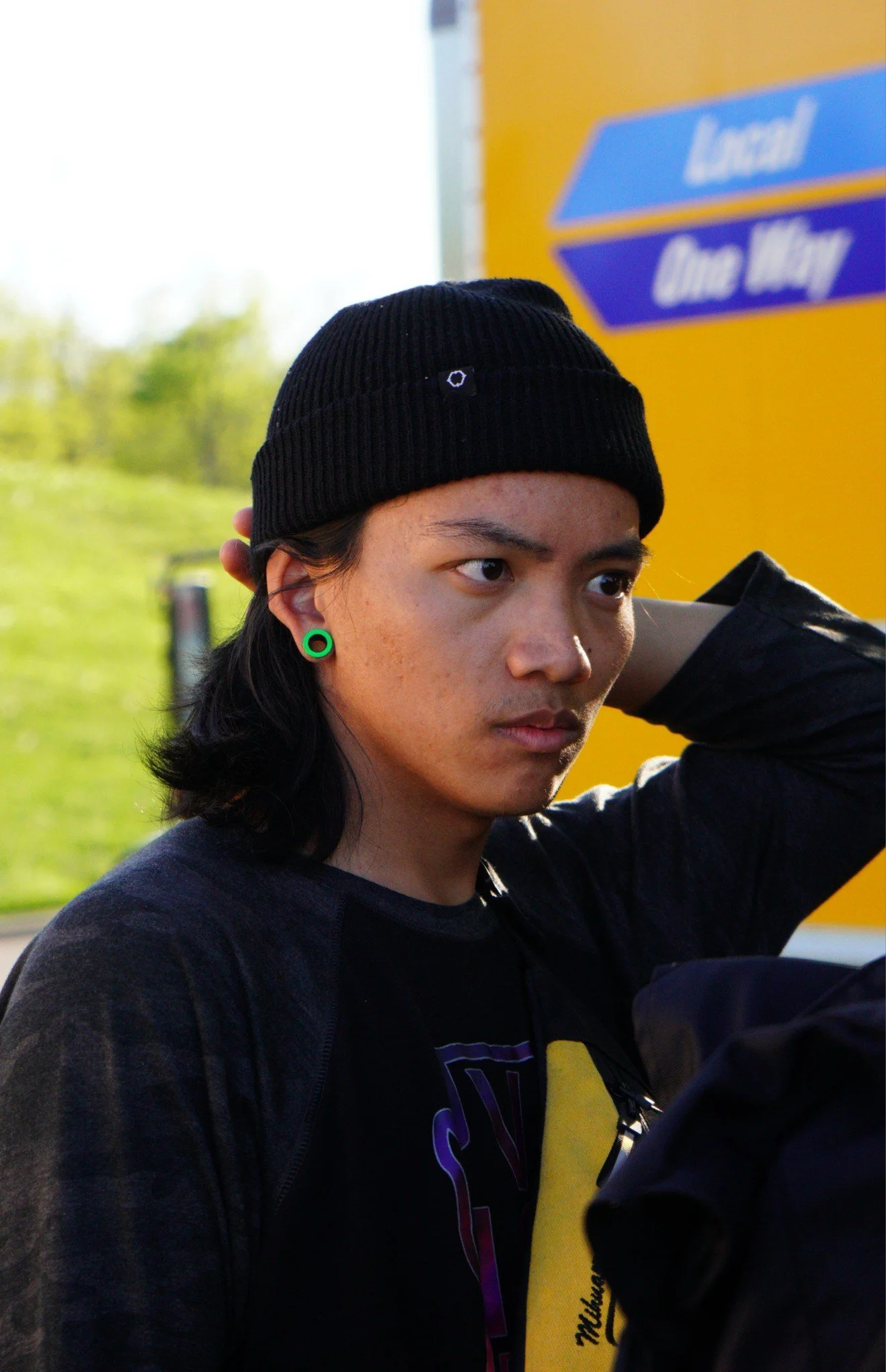 A young man with long black hair, wearing a black knit beanie and green gauge earrings, standing outdoors near a yellow sign with blue text, and looking serious with his hand behind his head.