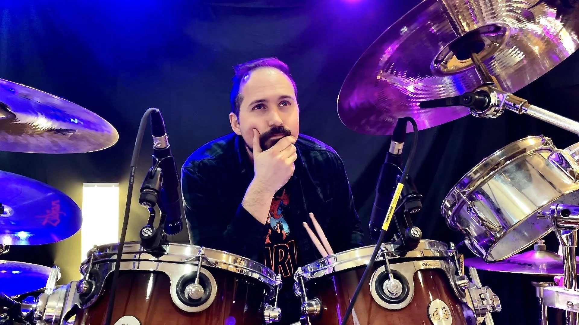 A man with a beard and mustache, wearing a black jacket and a Spider-Man t-shirt, is sitting behind a drum set with his hand on his chin, surrounded by drum cymbals and drums with microphones set up.