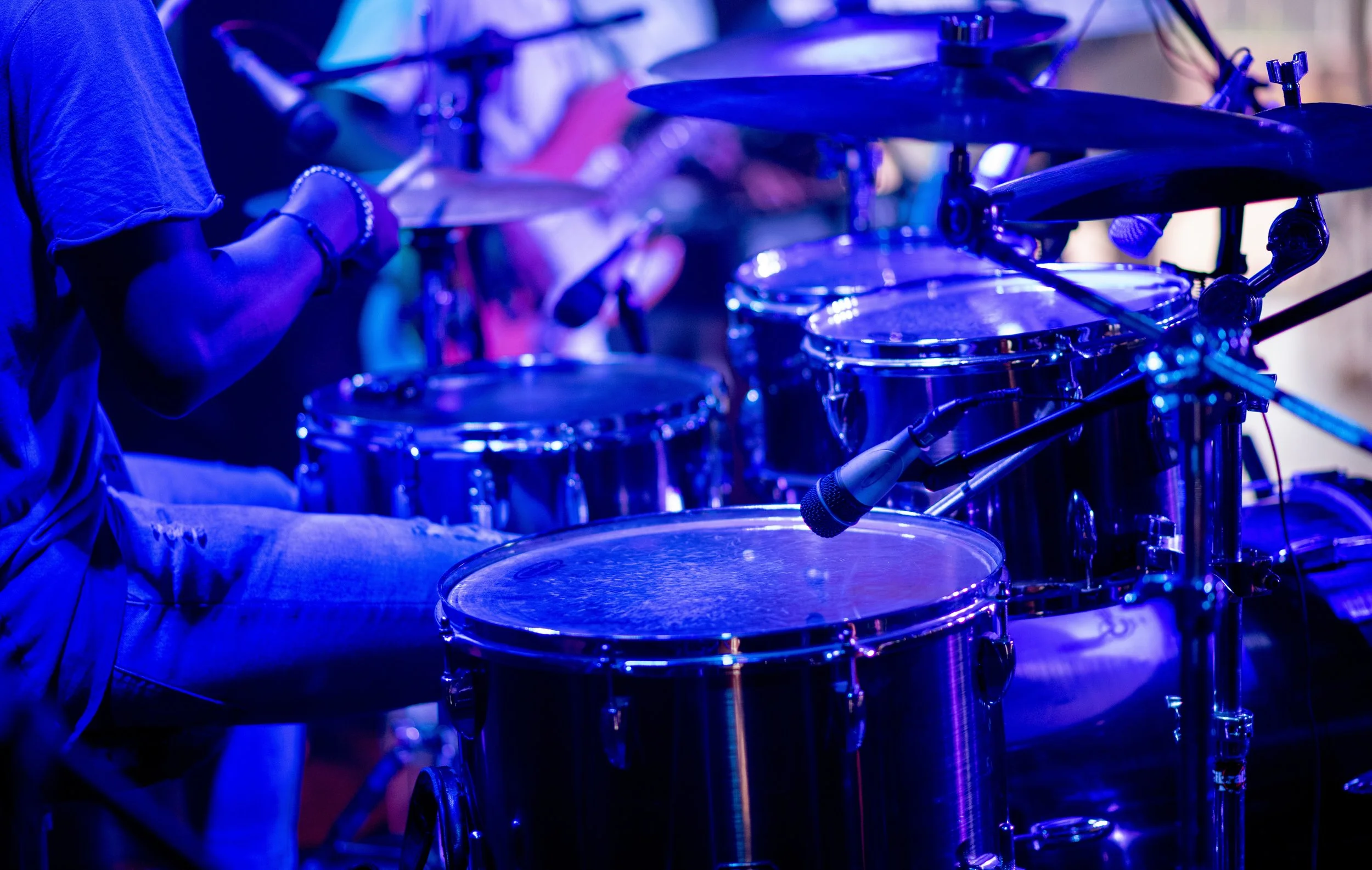 Close-up of a drum set being played by a person, with vivid blue lighting.