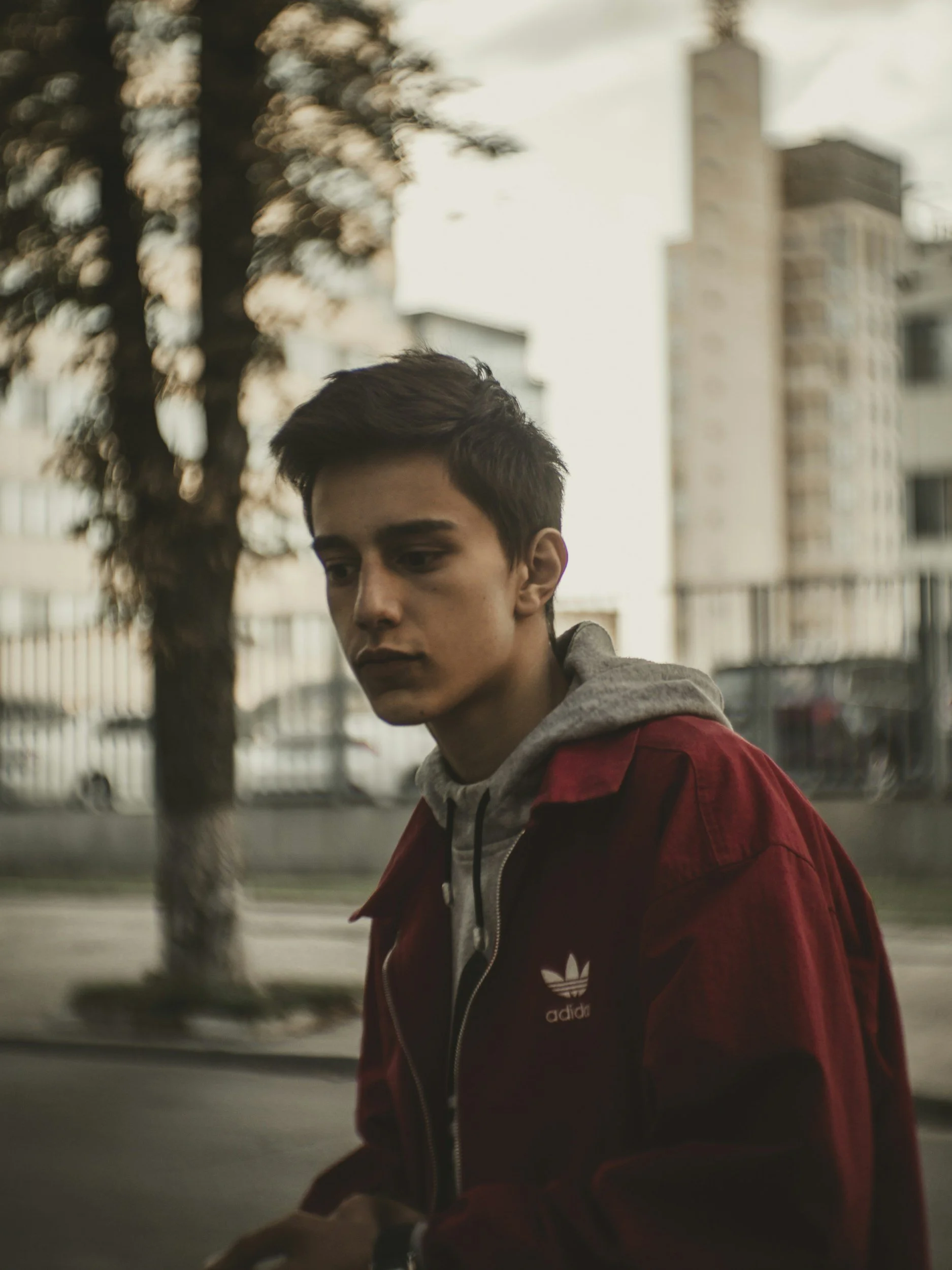 A teenage boy with brown hair and a red jacket on a city street looks down, contemplating his school stressors and low moods.