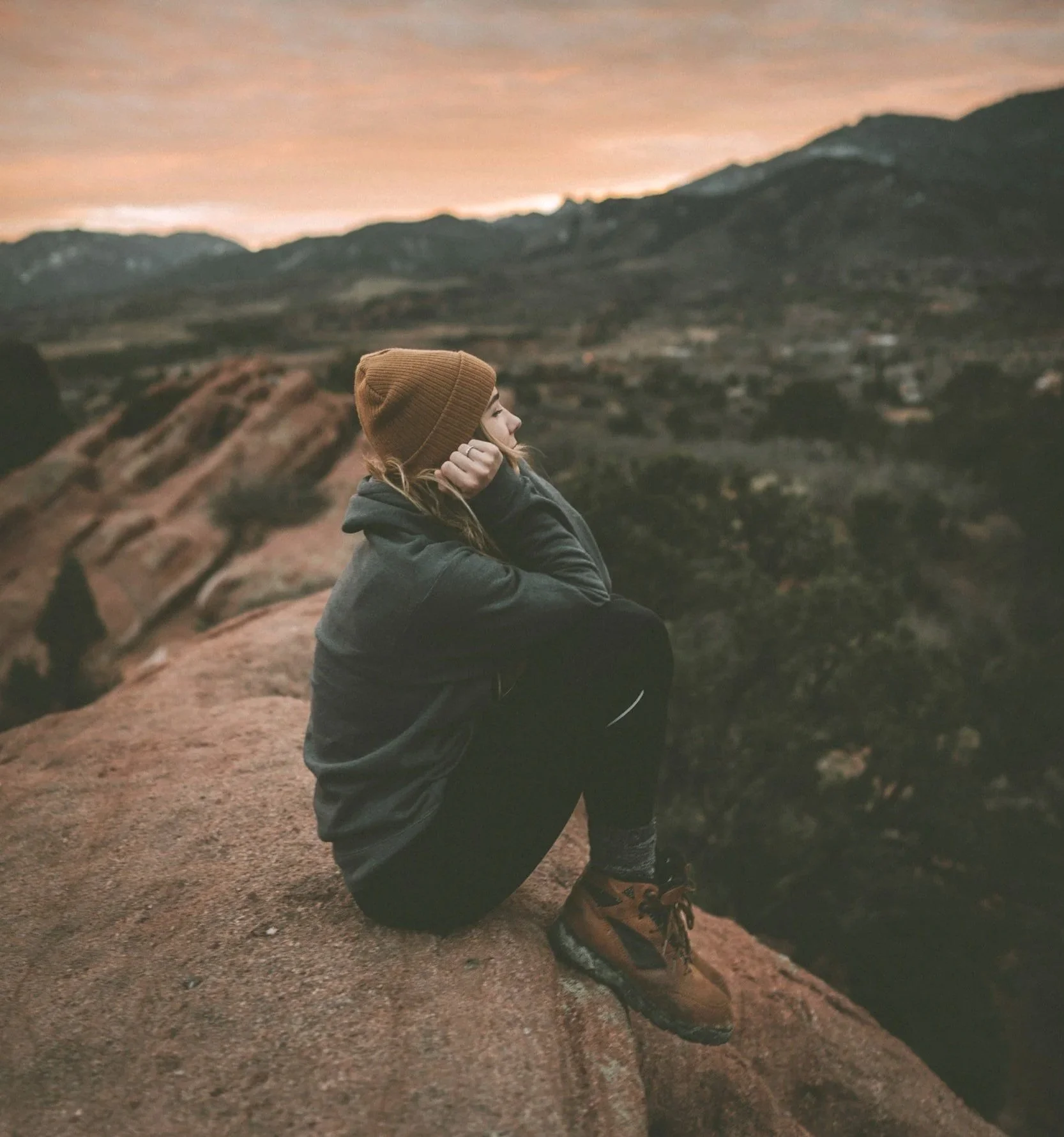 A young blonde woman wearing hiking apparel and a brown cap sits on a rock ledge, contemplating her mental health and wellness journey ahead. wellness.