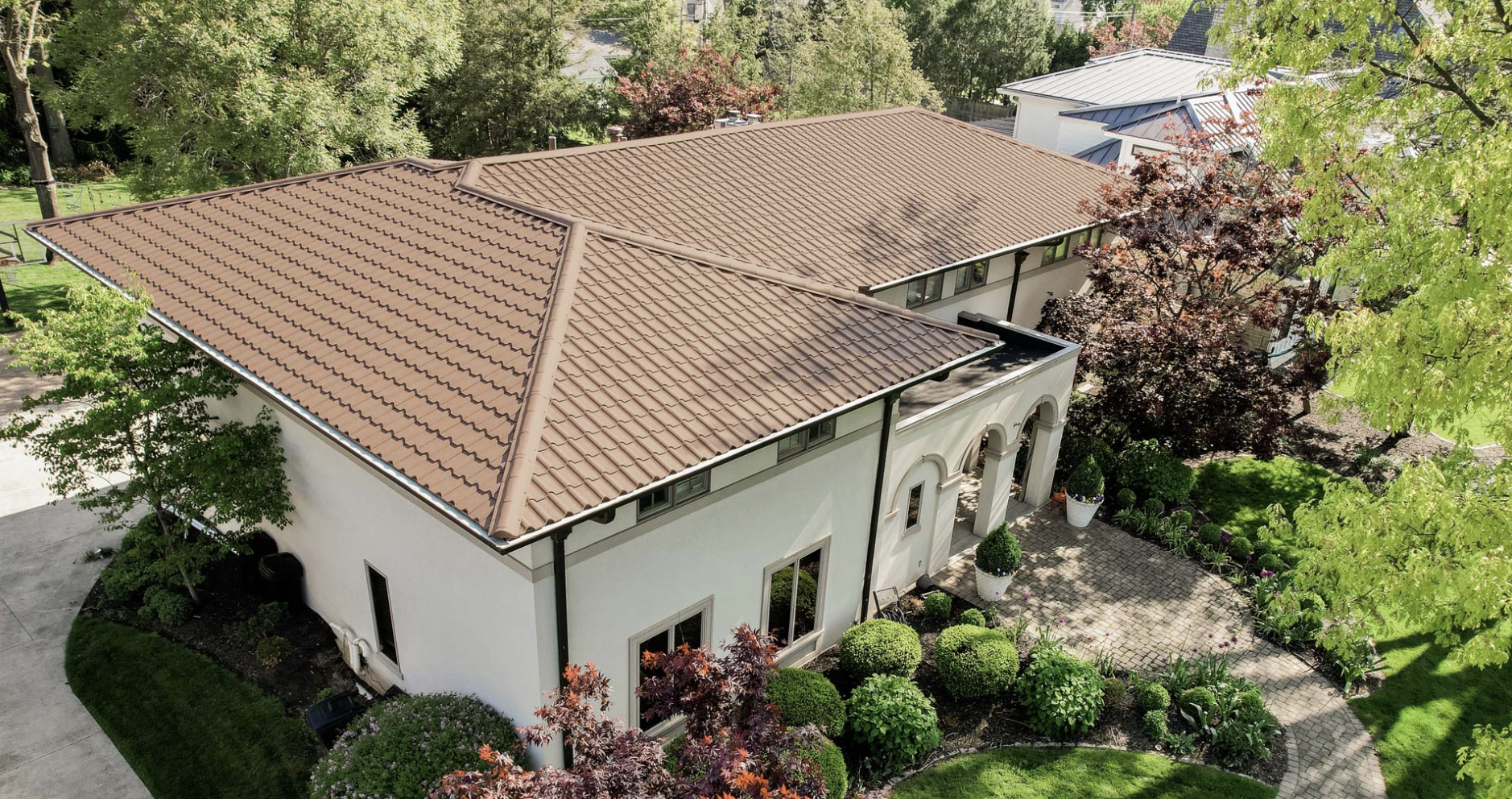 Aerial view of a white house with a brown tiled roof, surrounded by green trees and a landscaped garden with a stone pathway.