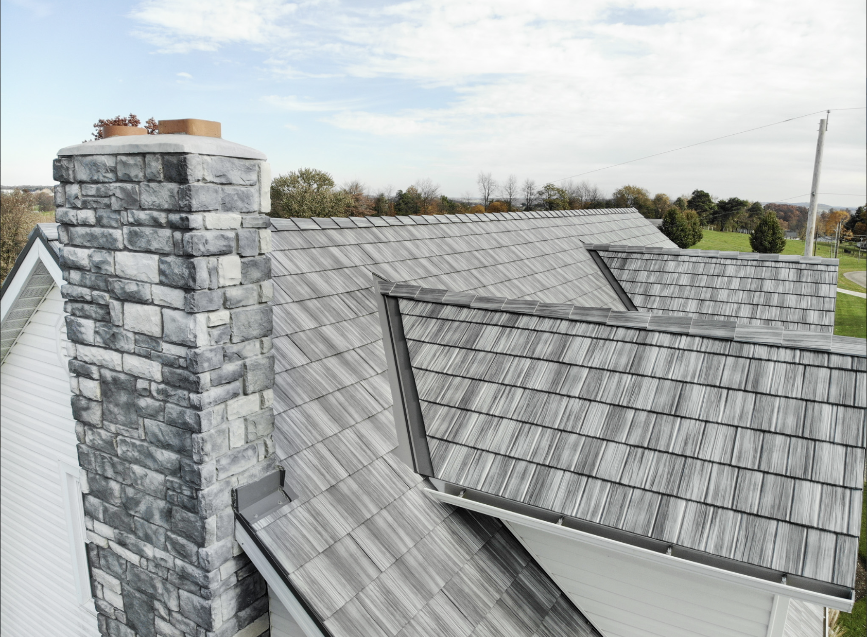 A house roof with gray shingles and a large stone chimney, with a rural landscape and partly cloudy sky in the background.