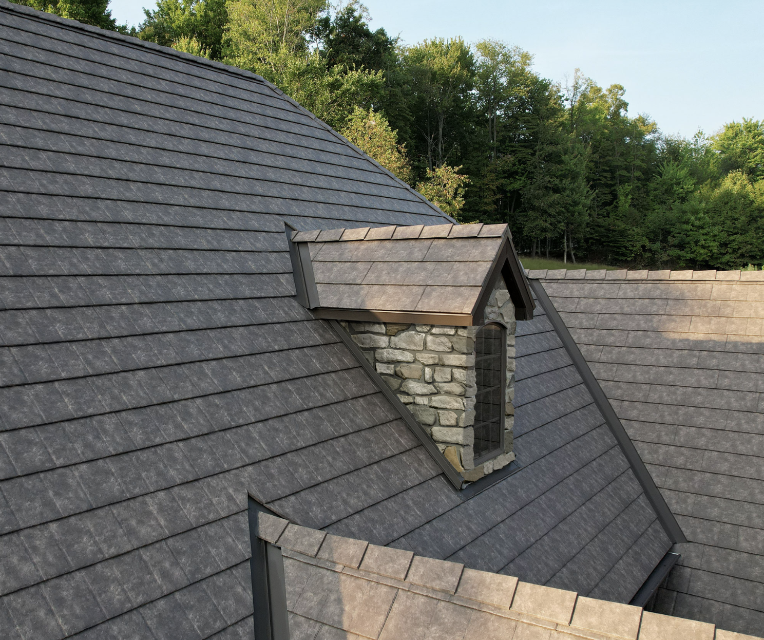 A roof with gray shingles and a stone chimney with a small gable roof on top, surrounded by green trees in the background.