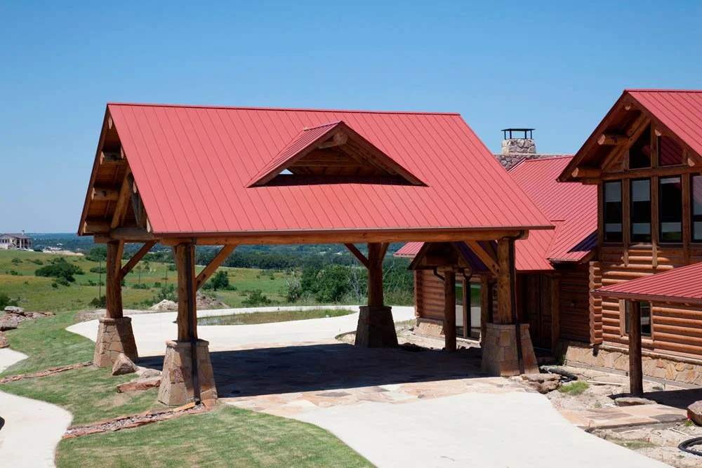 Wooden house with a red metal roof and a carport in a rural area with open fields and a clear blue sky.