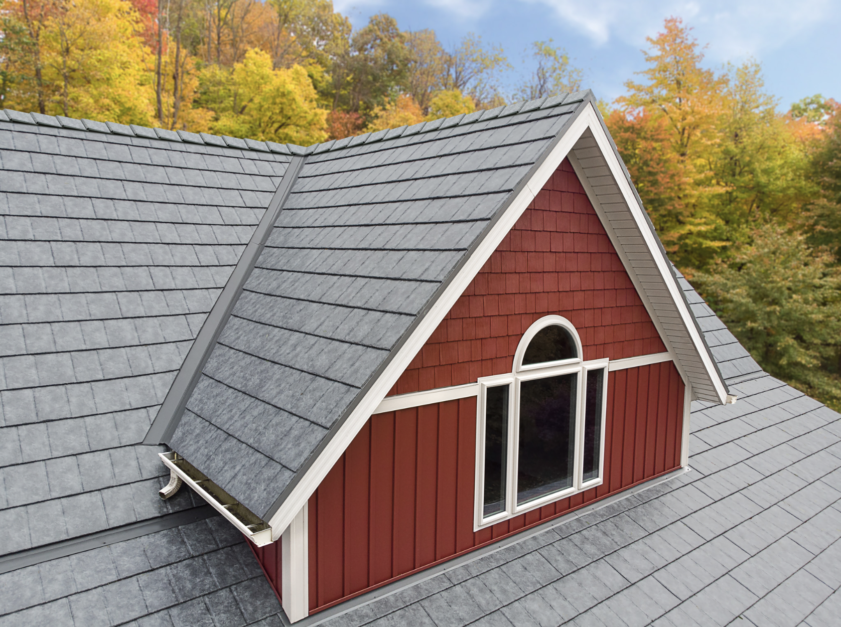 A red house with a gray shingled roof and a large arched window. The house is surrounded by trees with colorful autumn foliage.