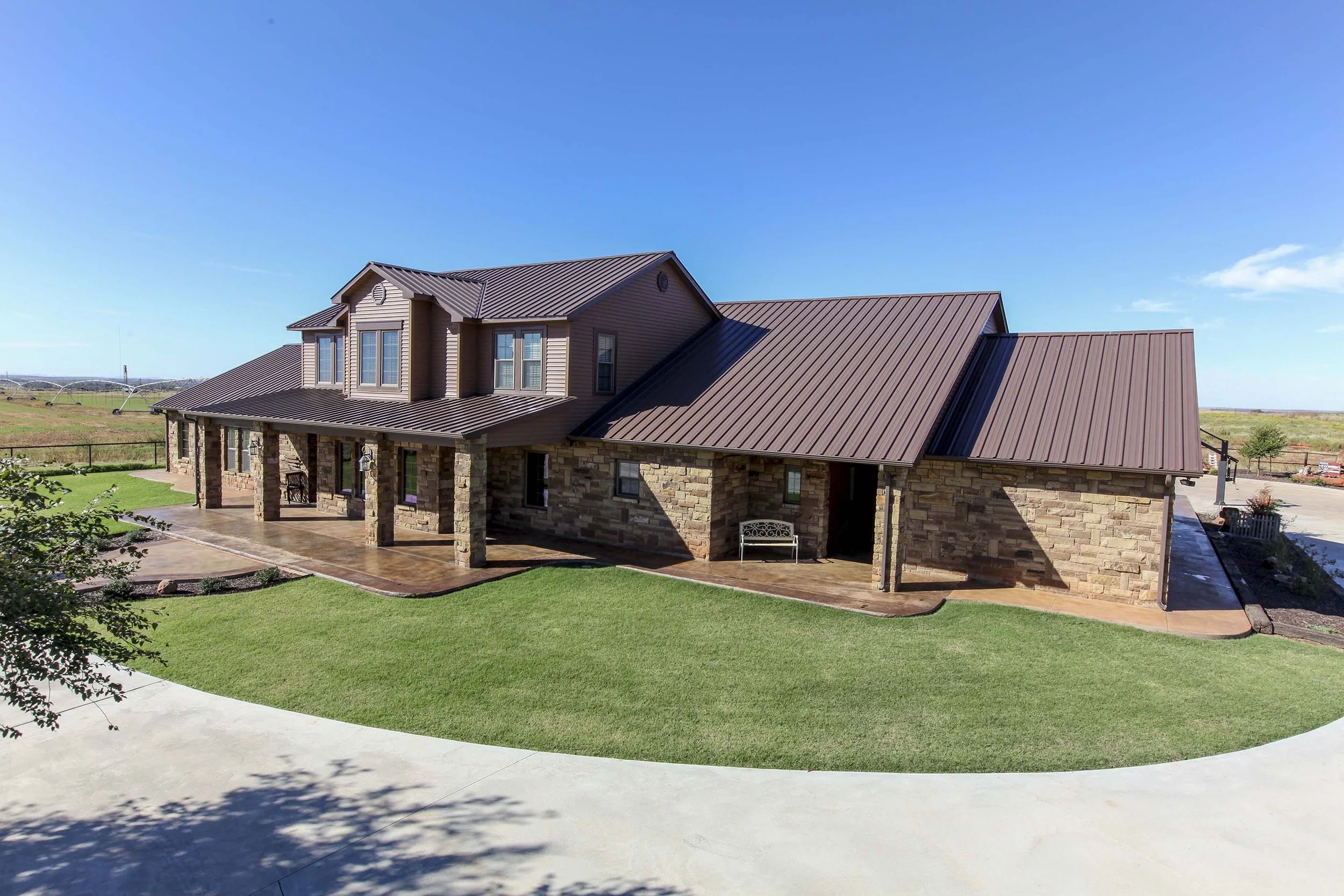 A large two-story house with a stone facade, metal roof, and covered porch, surrounded by a well-maintained lawn and open fields under a clear blue sky.