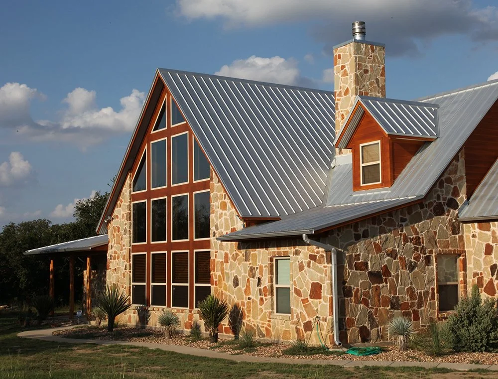 A house with a stone exterior, large front windows, a metal roof, and a chimney. The house has a small dormer window with a wooden frame, surrounded by landscaped yard with desert plants.