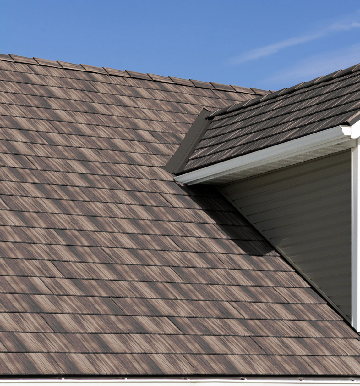 Close-up view of a house roof with wood-like shingles and a small section of grey siding, against a clear blue sky.