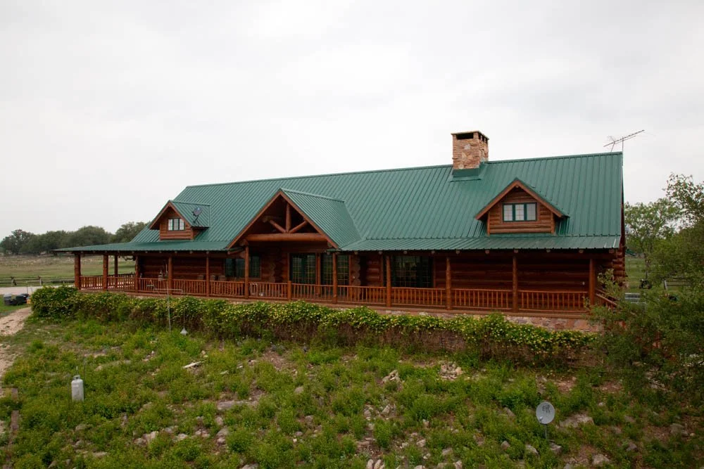 A large wooden house with a green metal roof, a brick chimney, and a covered front porch, situated on a grassy hill with a few trees in the background.