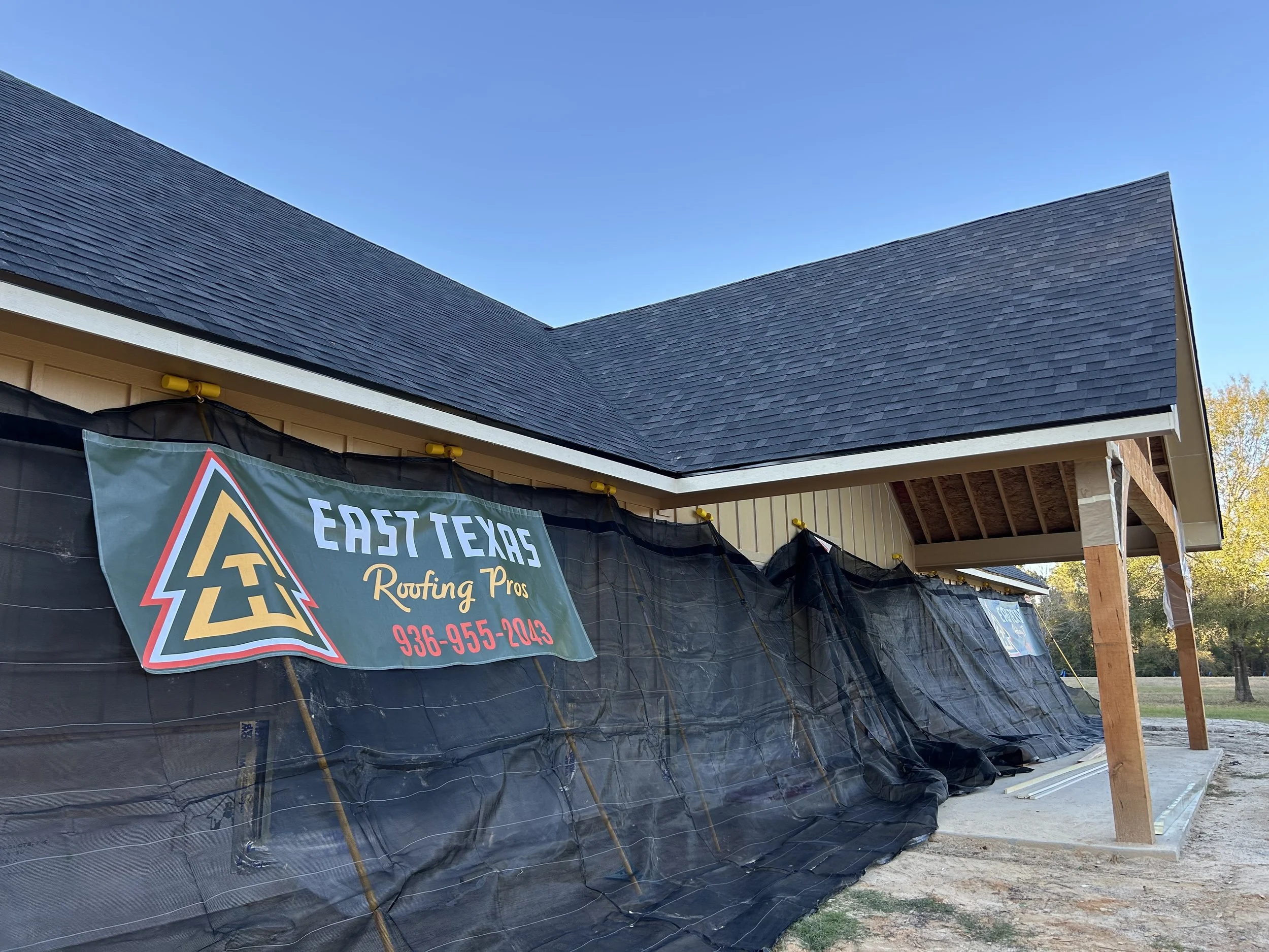 Construction site of a house with building materials and a sign for East Texas Routing Pros, showing the house's roof and exterior walls, with a black tarp covering part of the structure.