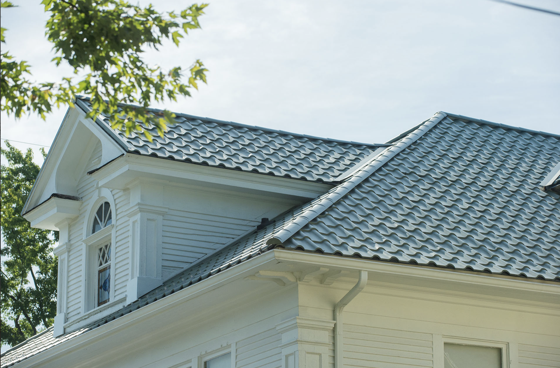 Close-up of a white house's roof with grey tiles, a dormer window with stained glass, and trees in the background.
