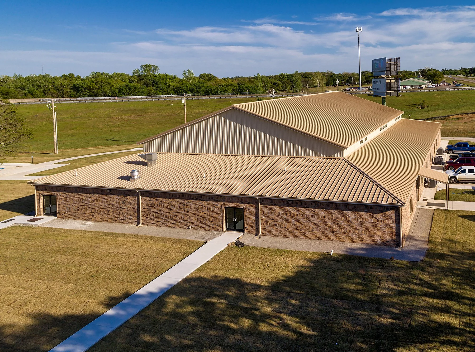 An aerial view of a single-story brick building with a beige metal roof, situated on a grassy area with sidewalks. In the background, there is a parking lot with several parked cars, utility poles, and a highway with billboards and a green landscape 