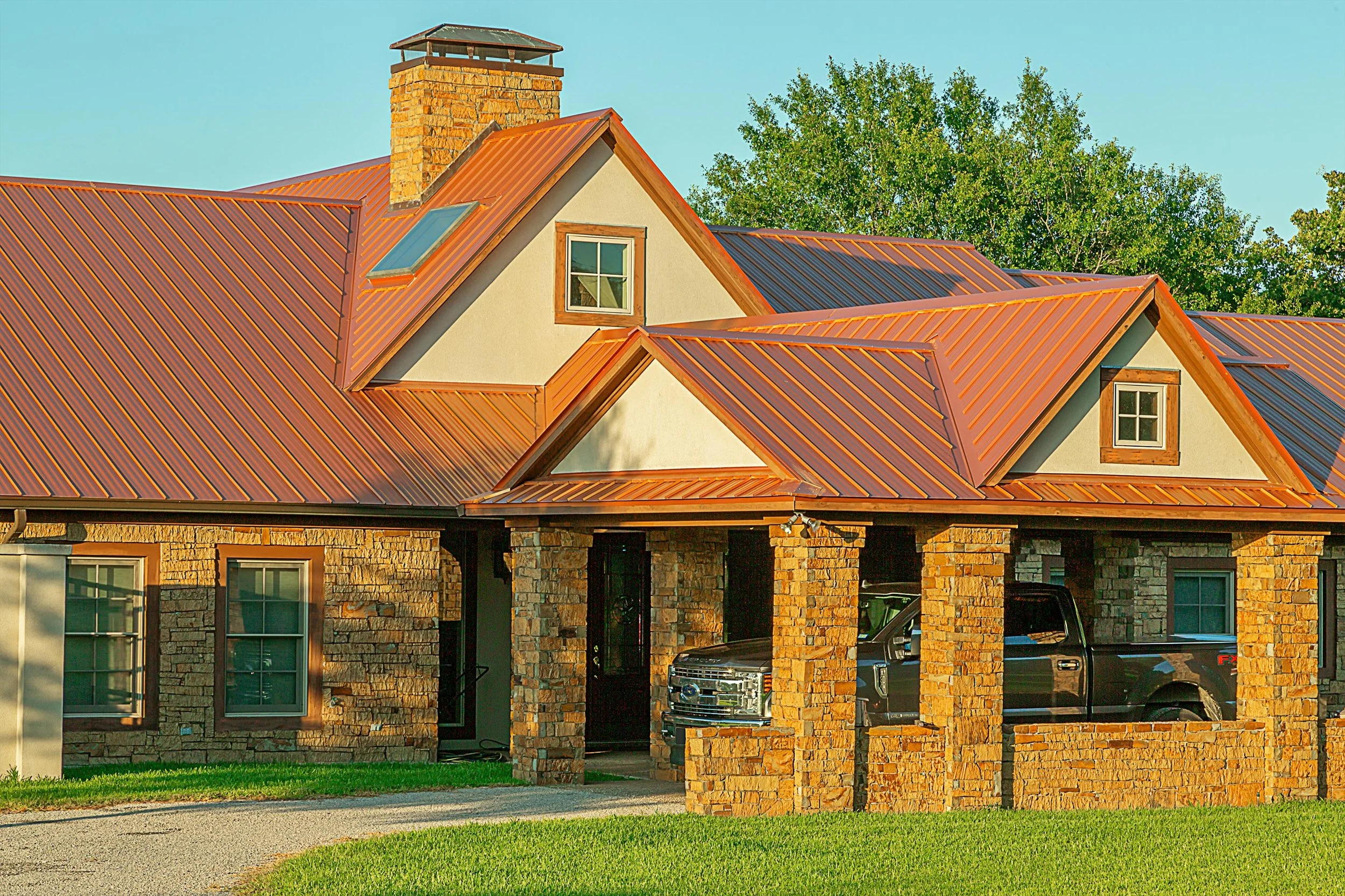 A house with a stone facade, multiple gabled roofs with copper-colored metal roofing, and a front porch with brick columns. Two trucks are parked inside the porch area, and there are double-hung windows visible on the house. Trees are in the backgrou