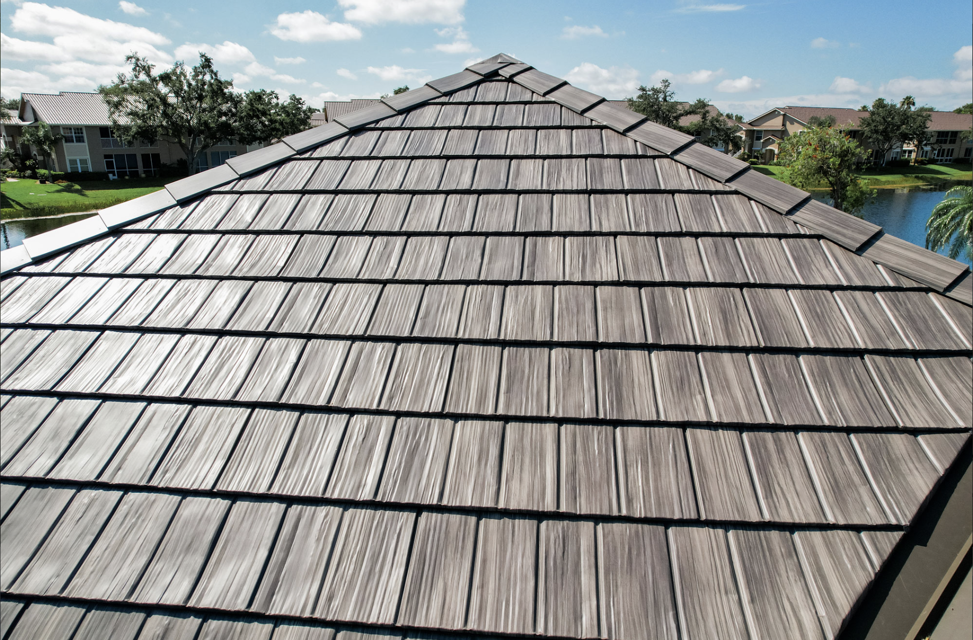 View of a brown tiled roof on a sunny day with a blue sky and scattered clouds, surrounded by green trees and residential buildings.