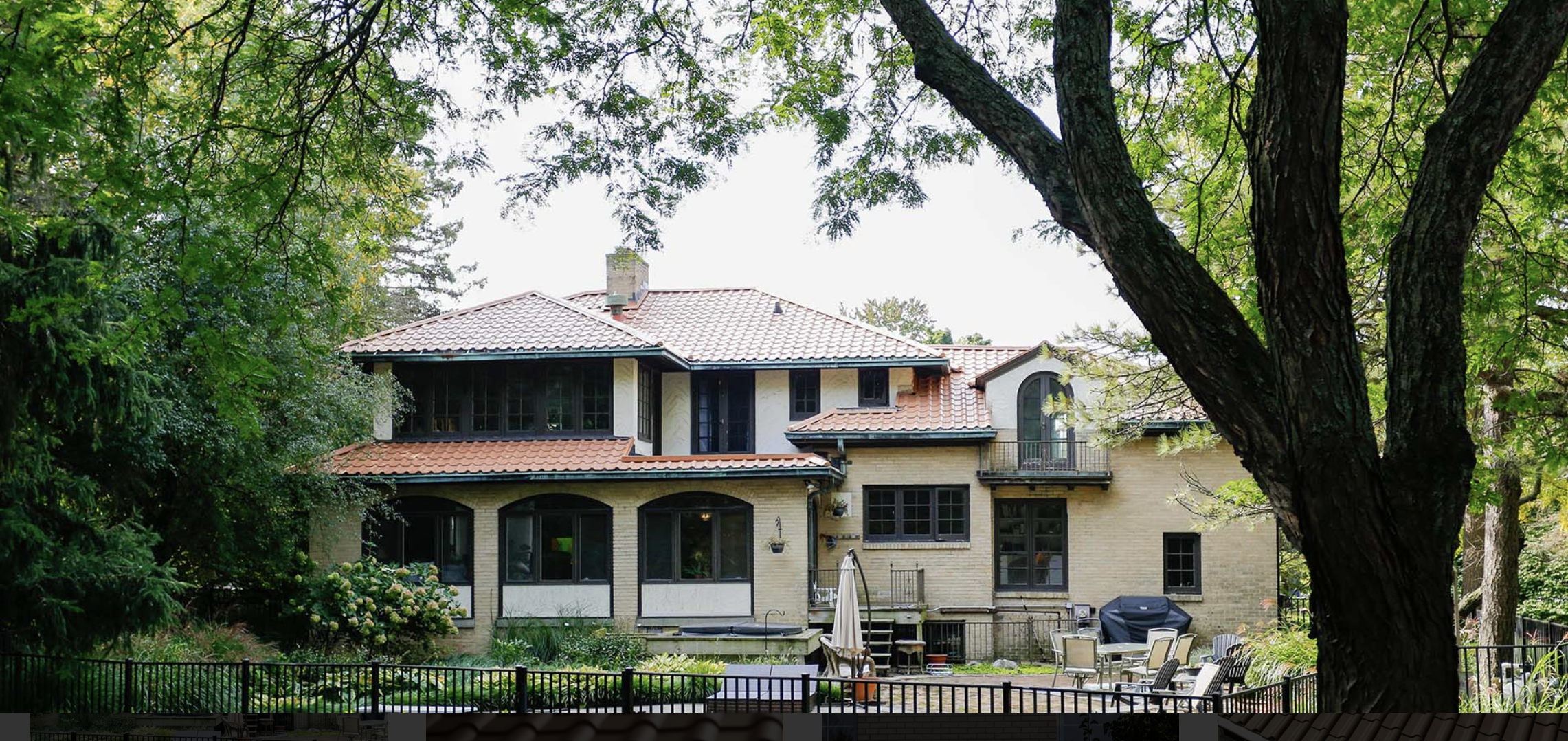 Backyard view of a large two-story house with a red tiled roof, surrounded by trees and greenery, with patio furniture and a black fence in the foreground.