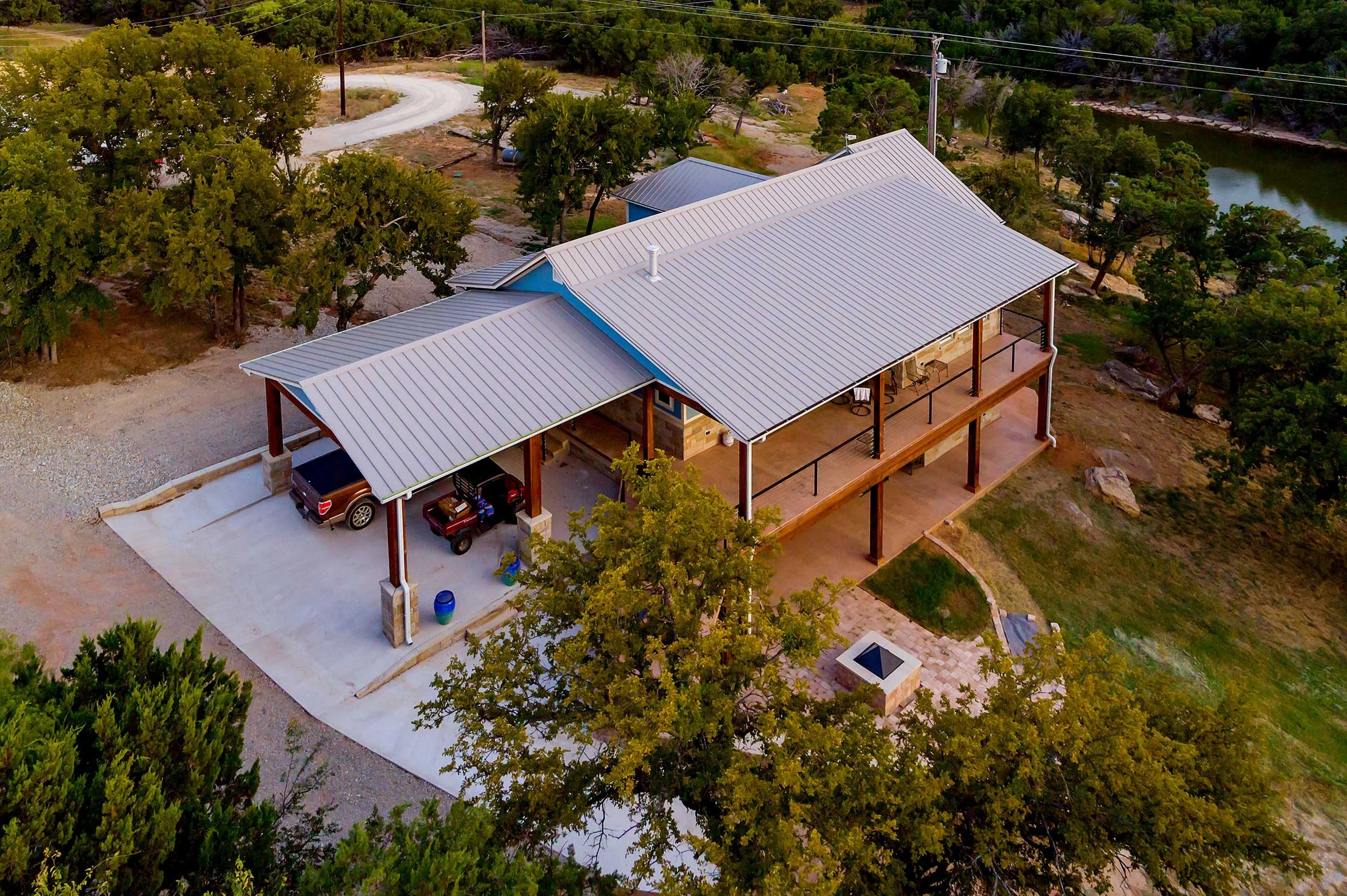 Aerial view of a house with a metal roof, a covered porch, and a driveway with two parked cars, surrounded by trees and a river in the background.