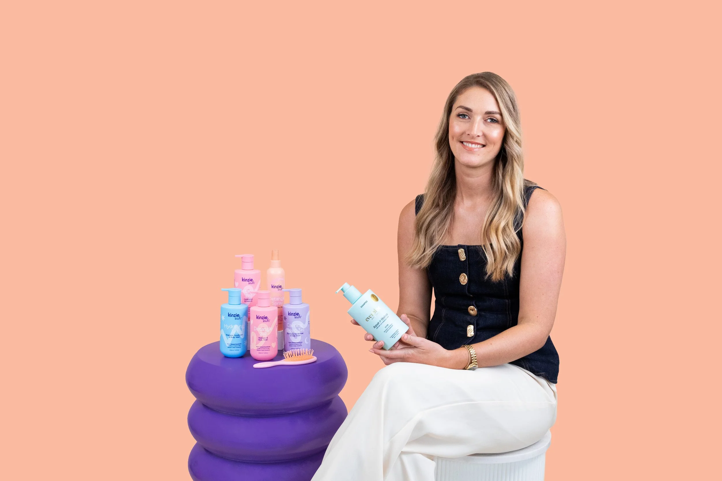 A woman sitting next to a purple stacked stool, holding a bottle of skincare product. Several more haircare bottles and a pink hairbrush are on the stool against a peach background.