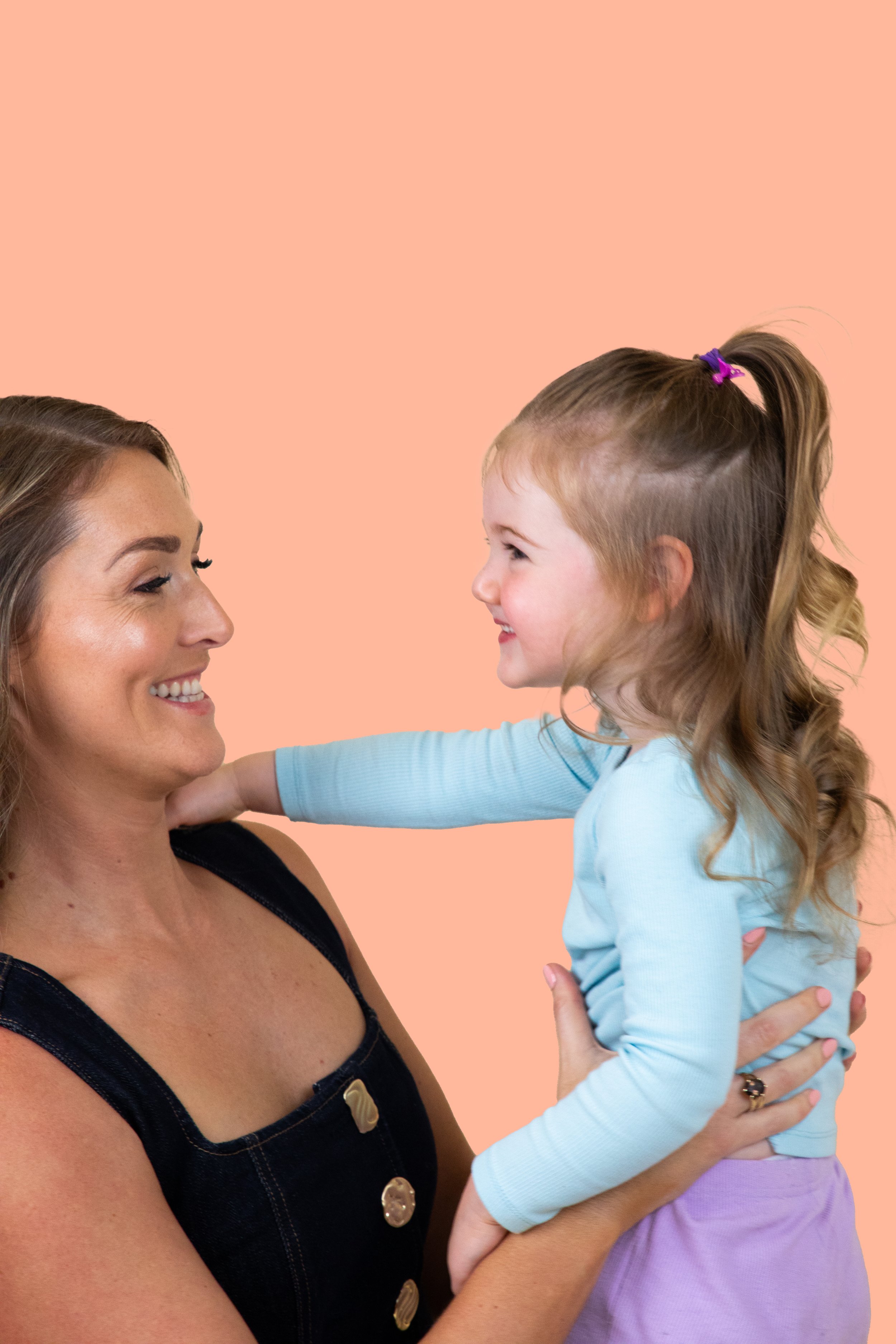 A woman holding a young girl in her arms, both smiling and looking at each other, against a peach-colored background.
