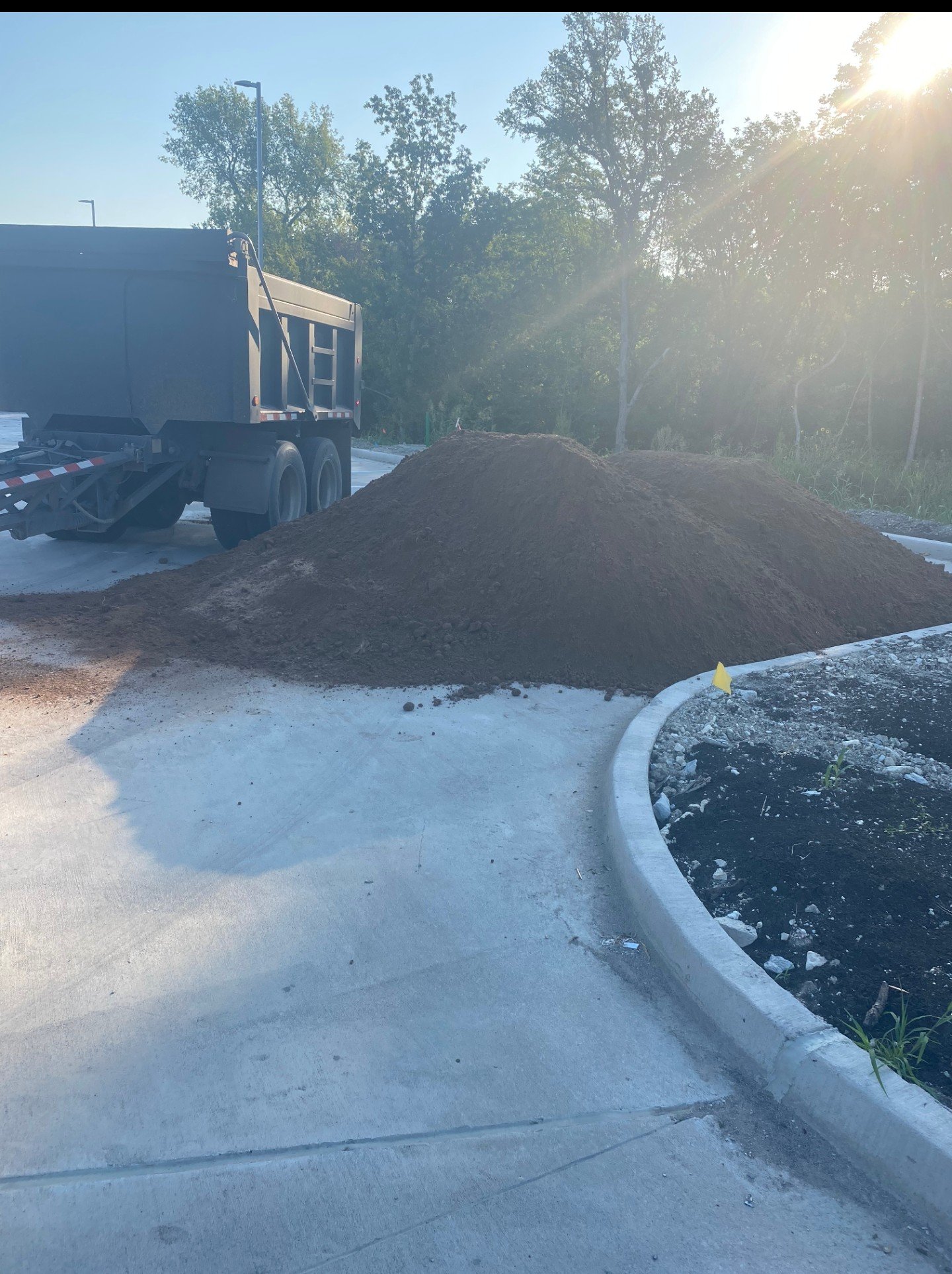 A pile of dirt beside a curb on a paved surface, with a dump truck parked nearby. The scene is outdoors during daylight, with trees and sunlight in the background.