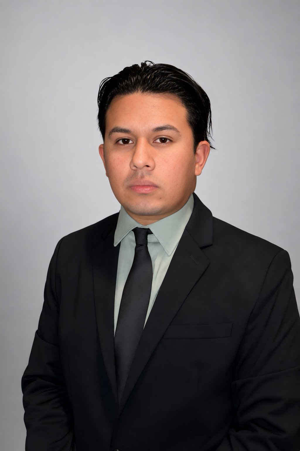 A man in a black suit with a light shirt and black tie, looking directly at the camera against a plain gray background.